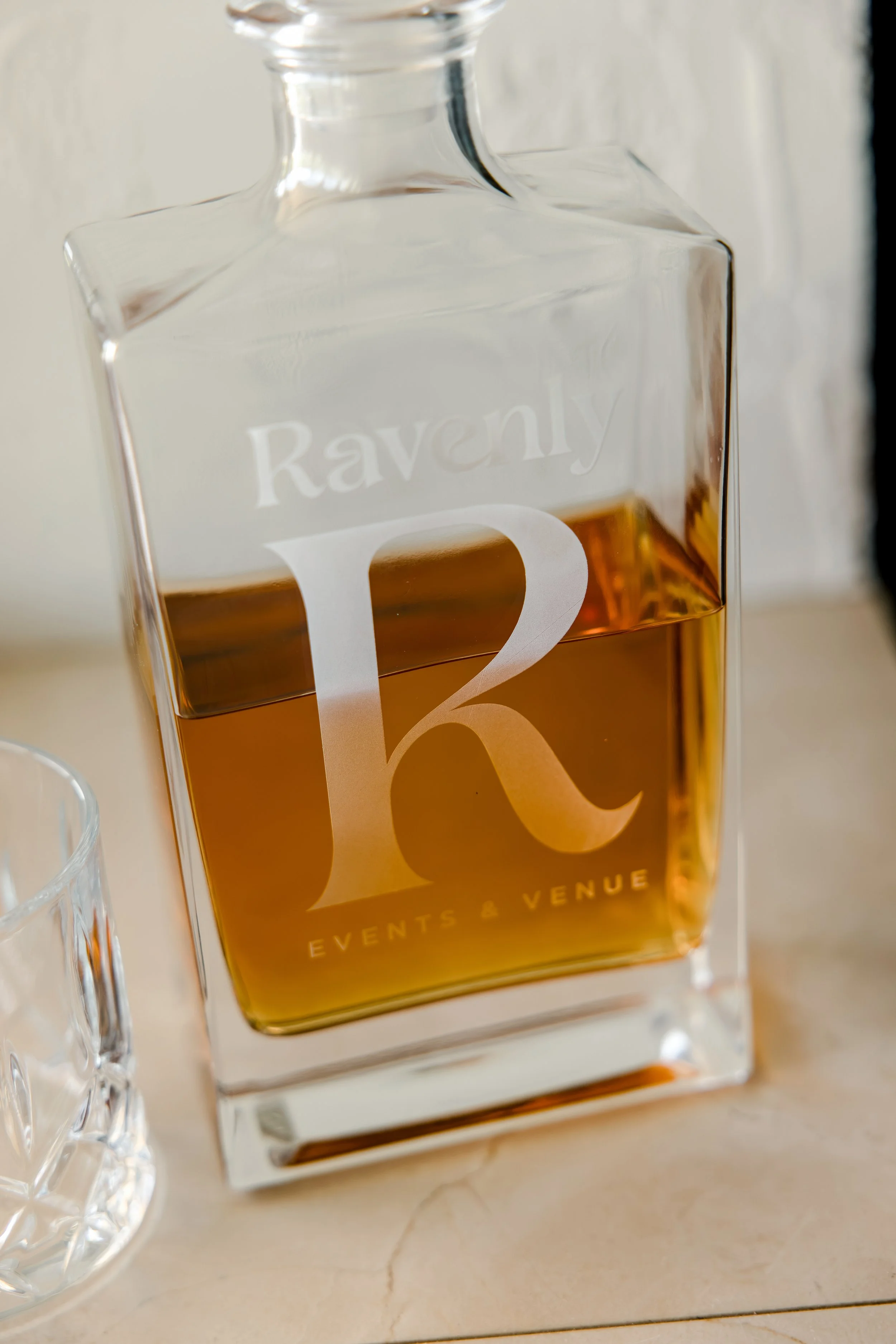A clear glass decanter filled with amber-colored liquor, with the word 'Raveny' and the letter 'K' visible on the front, placed on a light-colored marble or stone surface.