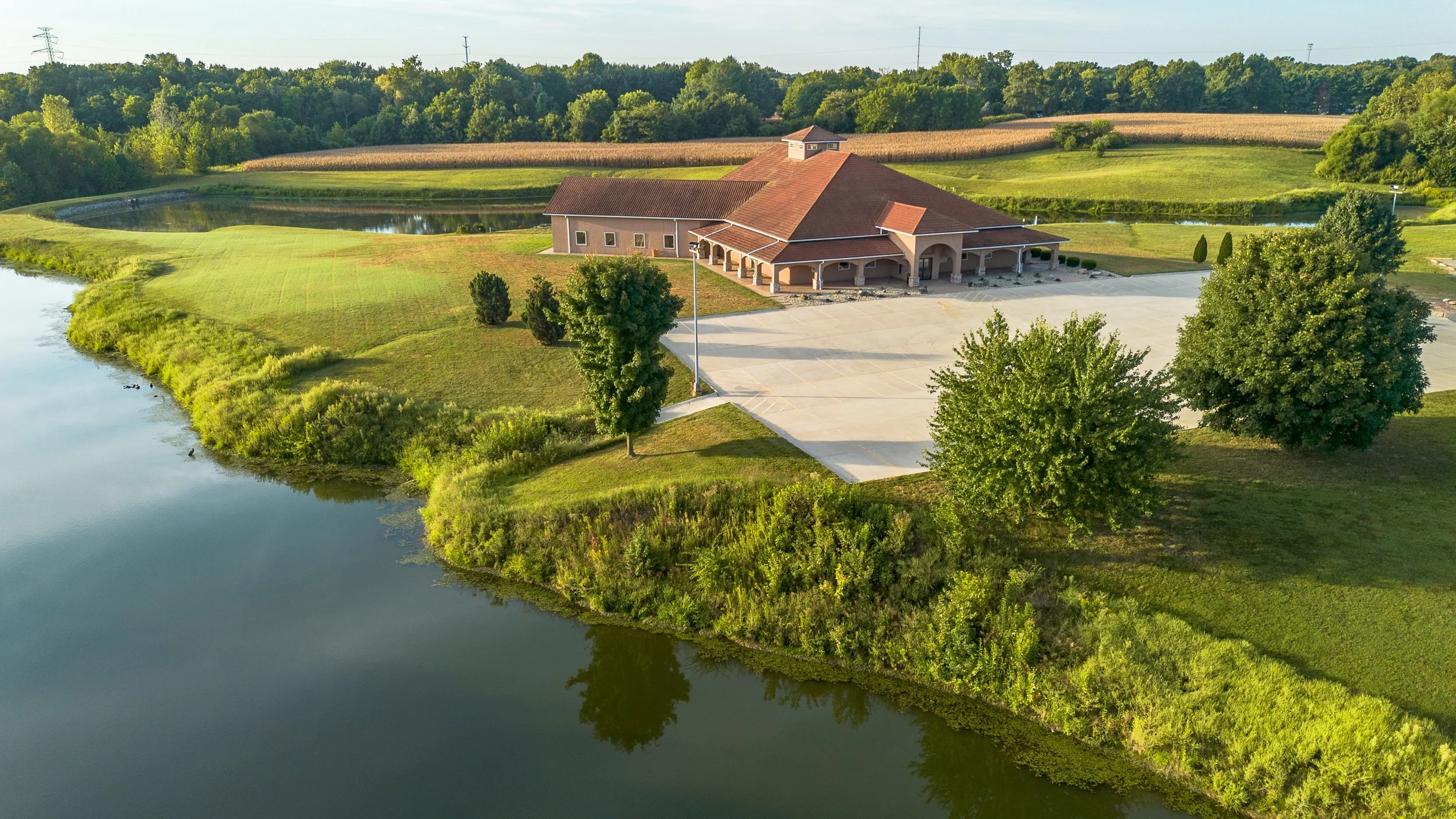 A church surrounded by green grass, trees, and a pond with ducks, with an expansive parking lot in front.