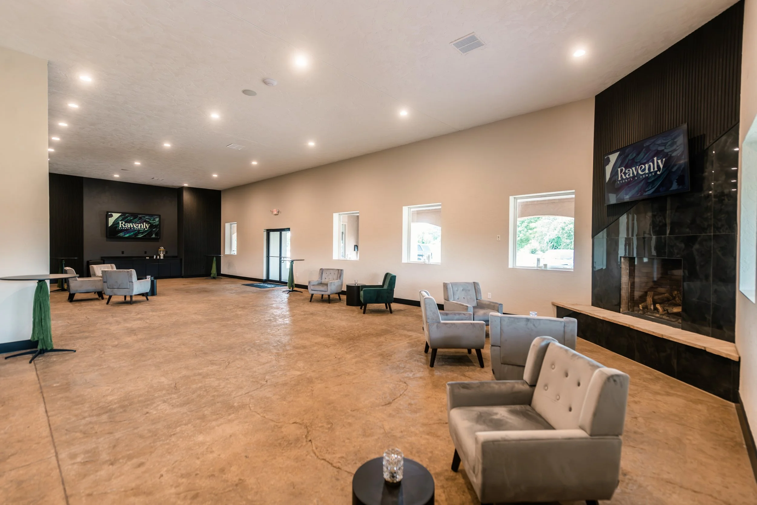 A spacious event room with multiple seating areas, windows, and two wall-mounted screens displaying 'Ravenly.' There is a fireplace on the right side and a few small tables with chairs around the room.