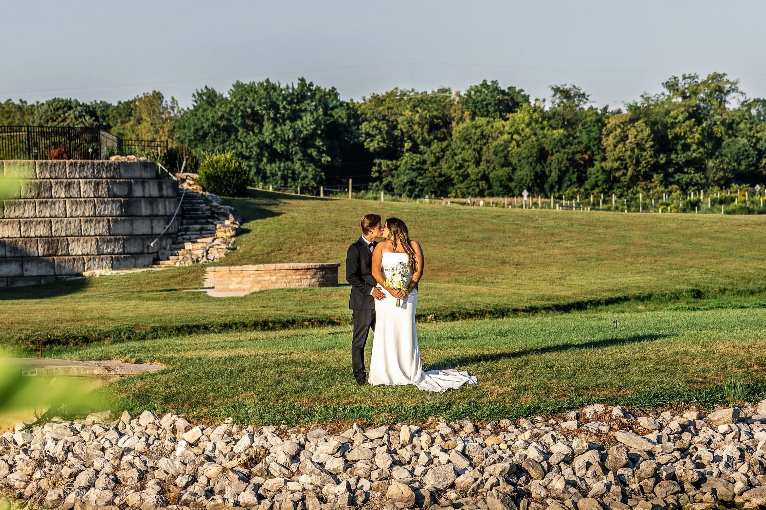 A couple in wedding attire standing on a grassy area outdoors, sharing a kiss, with lush green trees in the background.