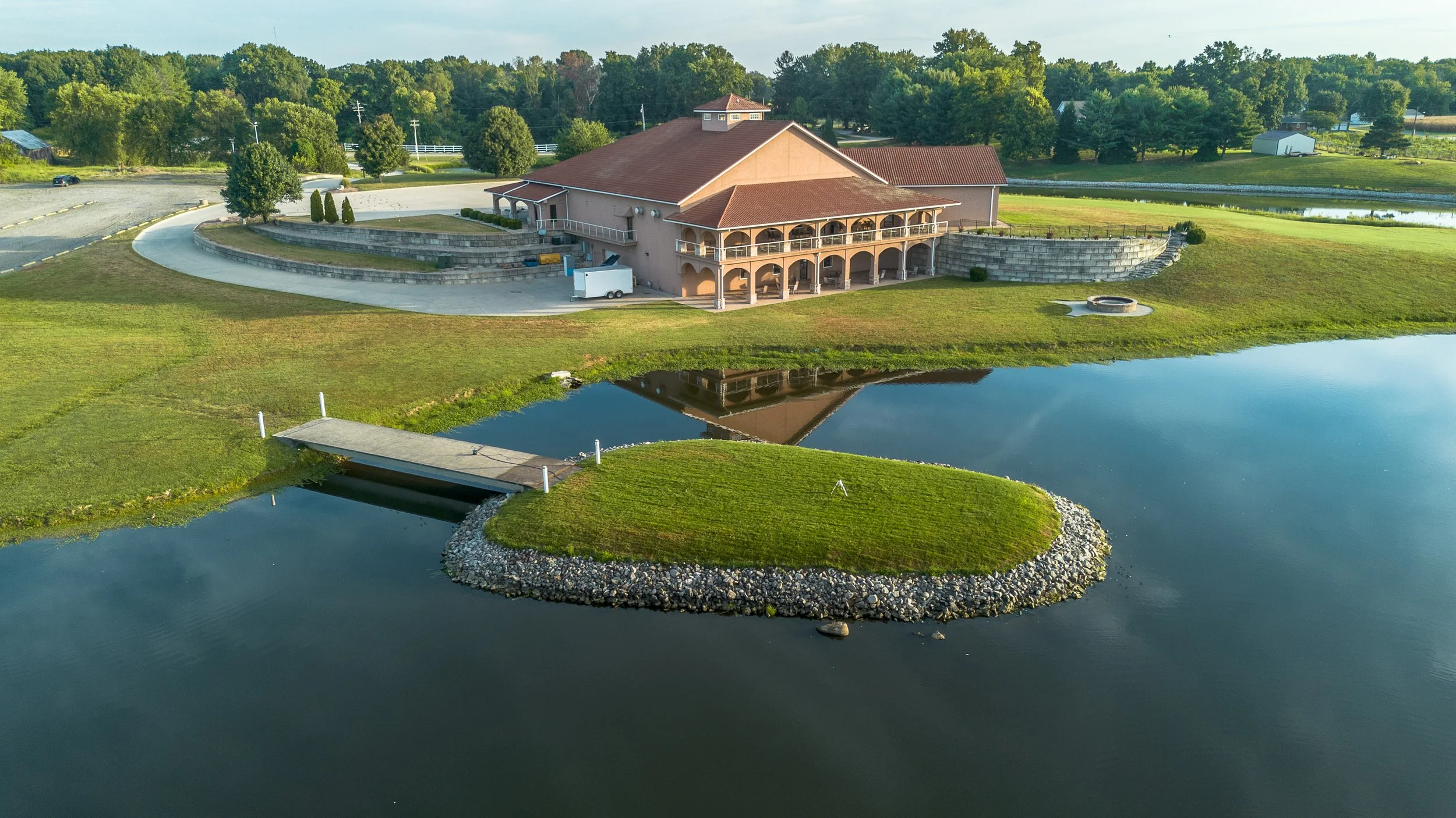 A large building with pinkish walls and red-tiled roof is situated by a pond, with a small island and a dock in the water, surrounded by green grass, trees, and a parking lot in the background.