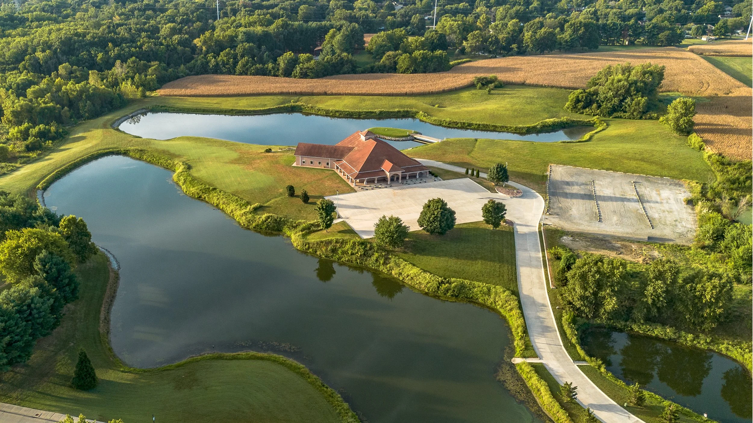 An aerial view of a large estate with two ponds, a main building with a red roof, a parking lot, and surrounding trees and fields.
