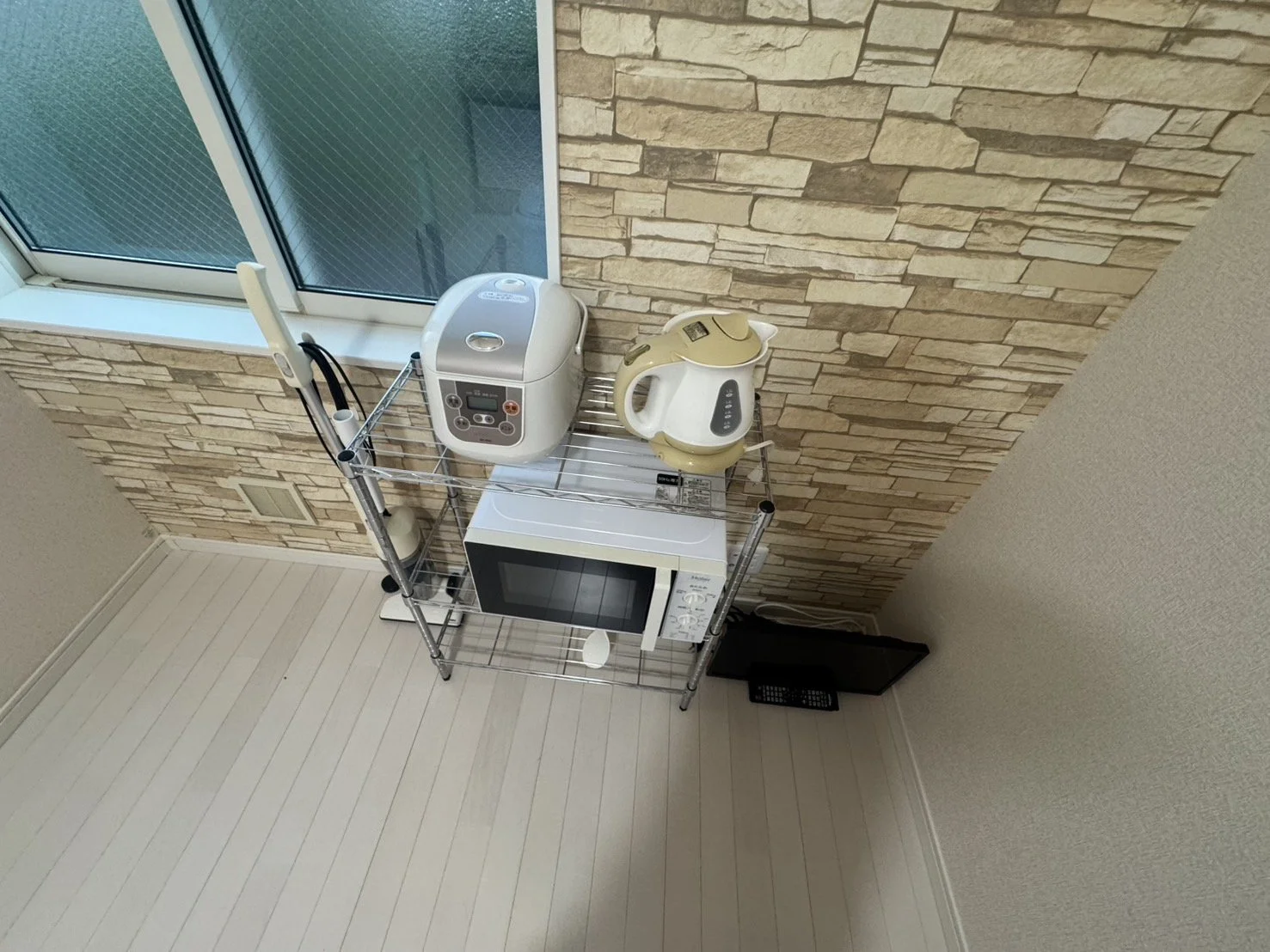 キッチンの一角に白いレンジ、ホワイトの電気ポット、炊飯器が置かれた金属製のラックがあり、その下に電子レンジがある。背景は窓と石張りの壁。