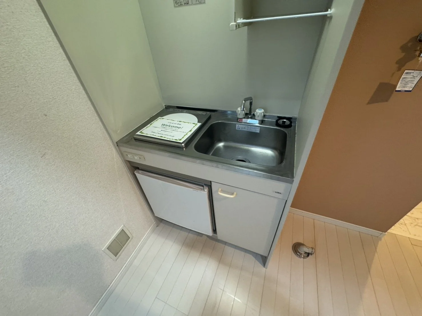 小さなキッチンシンクと冷蔵庫のついた簡易キッチン。ステンレス製のシンクには蛇口と缶詰が置かれ、上には紙の案内板があります。