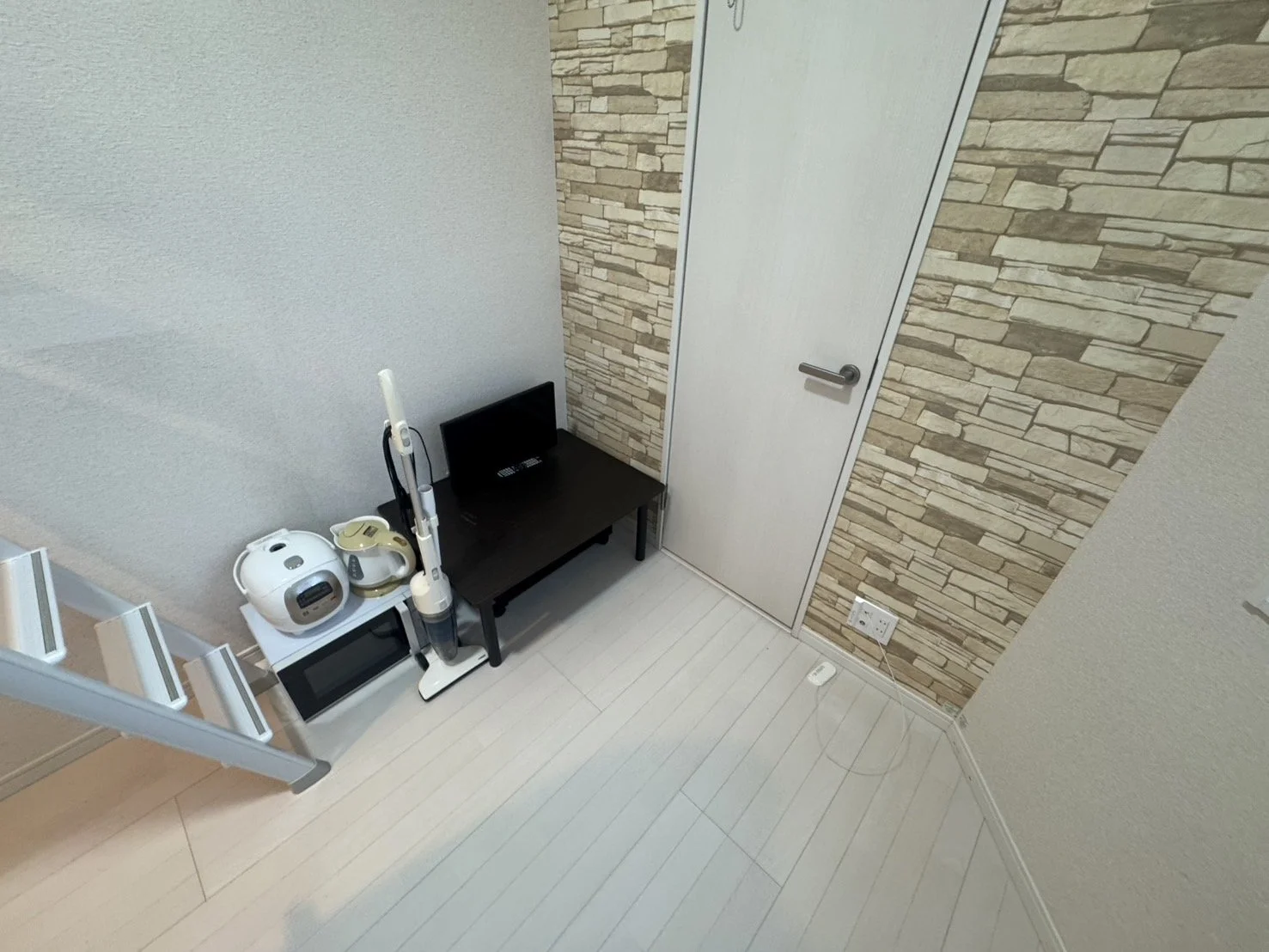 白と茶色のストーン調の壁紙と白いドアのある部屋に、ロボット掃除機と電話、テレビが置かれた黒いテーブル、電子レンジと炊飯器が棚に置いてある光沢のある白い床の部屋