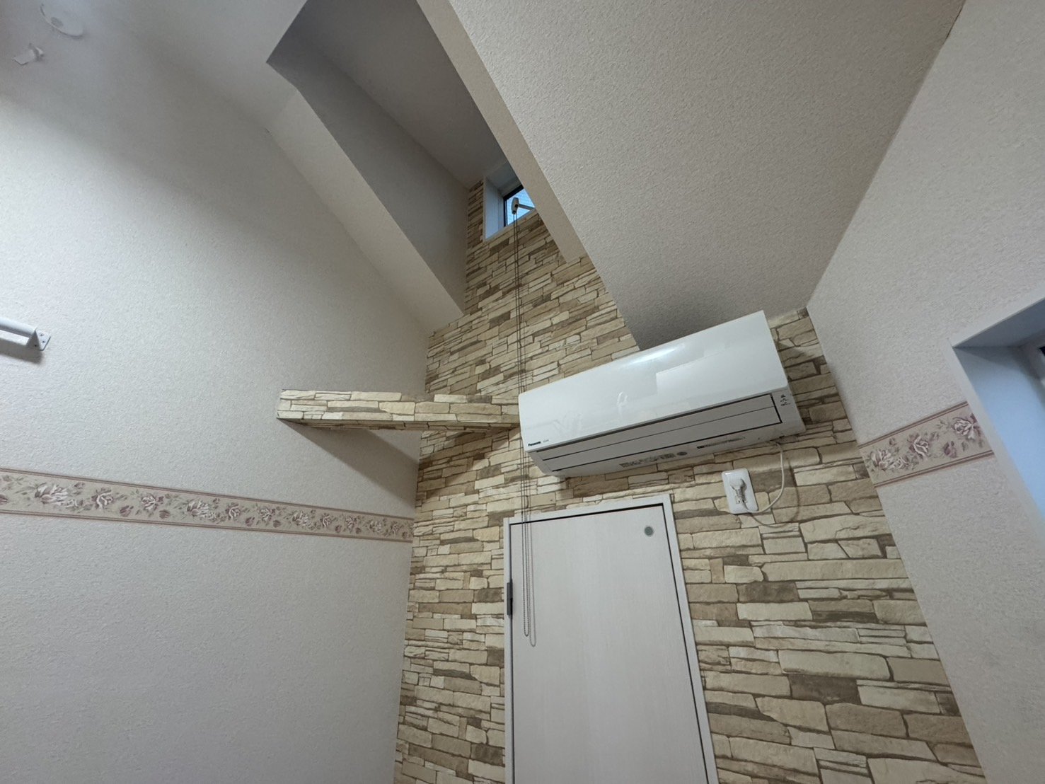 壁に取り付けられたエアコンと小さな窓、その周囲は石の壁とクロスの壁があり、天井も高い部屋の内装写真