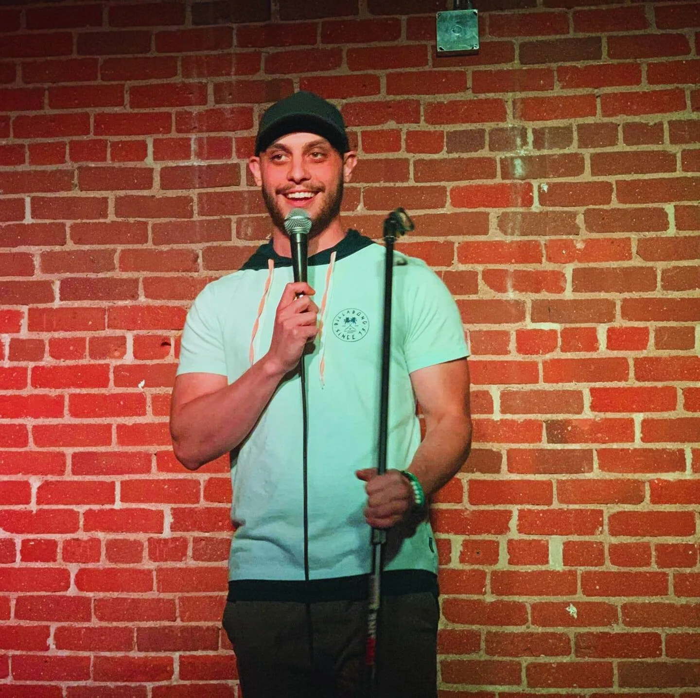 A man in a black beanie and light green T-shirt holding a microphone on stage with a brick wall background.