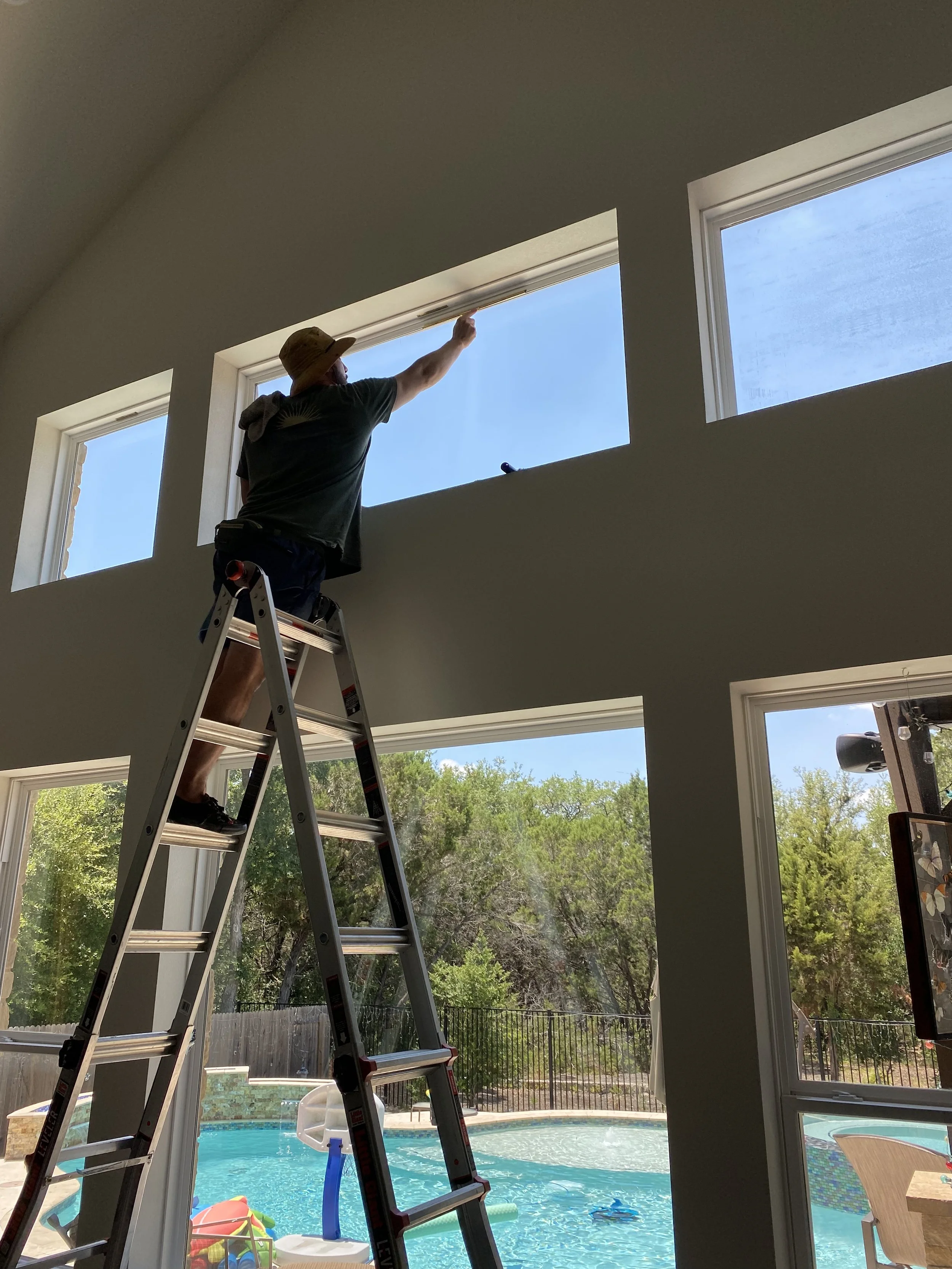 Man standing on a ladder, cleaning or installing a window in a high wall of a room, with a view of trees and a backyard with a swimming pool outside.