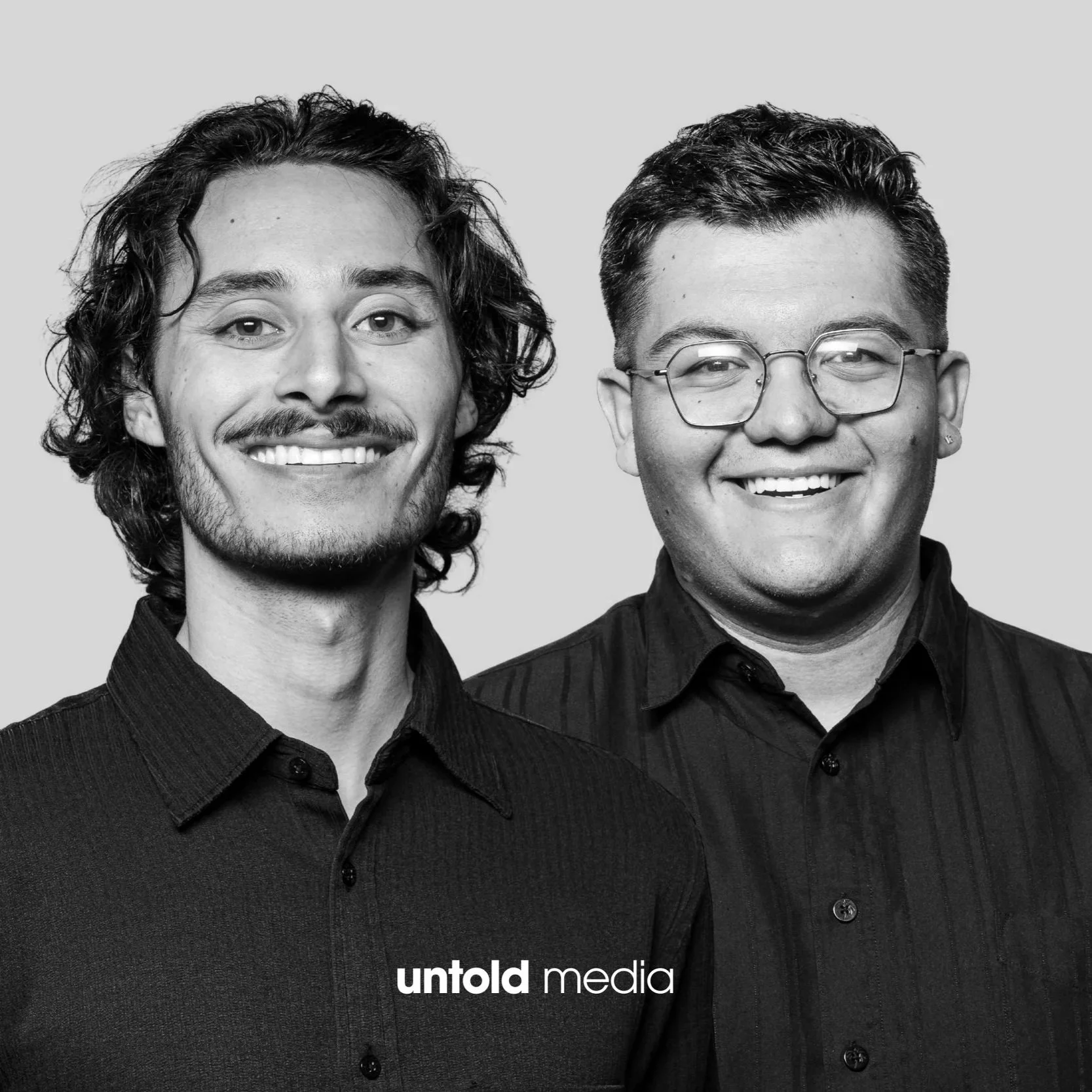 Black and white portrait of two smiling men wearing dark collared shirts, standing close together. The man on the left has curly hair and a mustache, while the man on the right is wearing glasses and has short hair.
