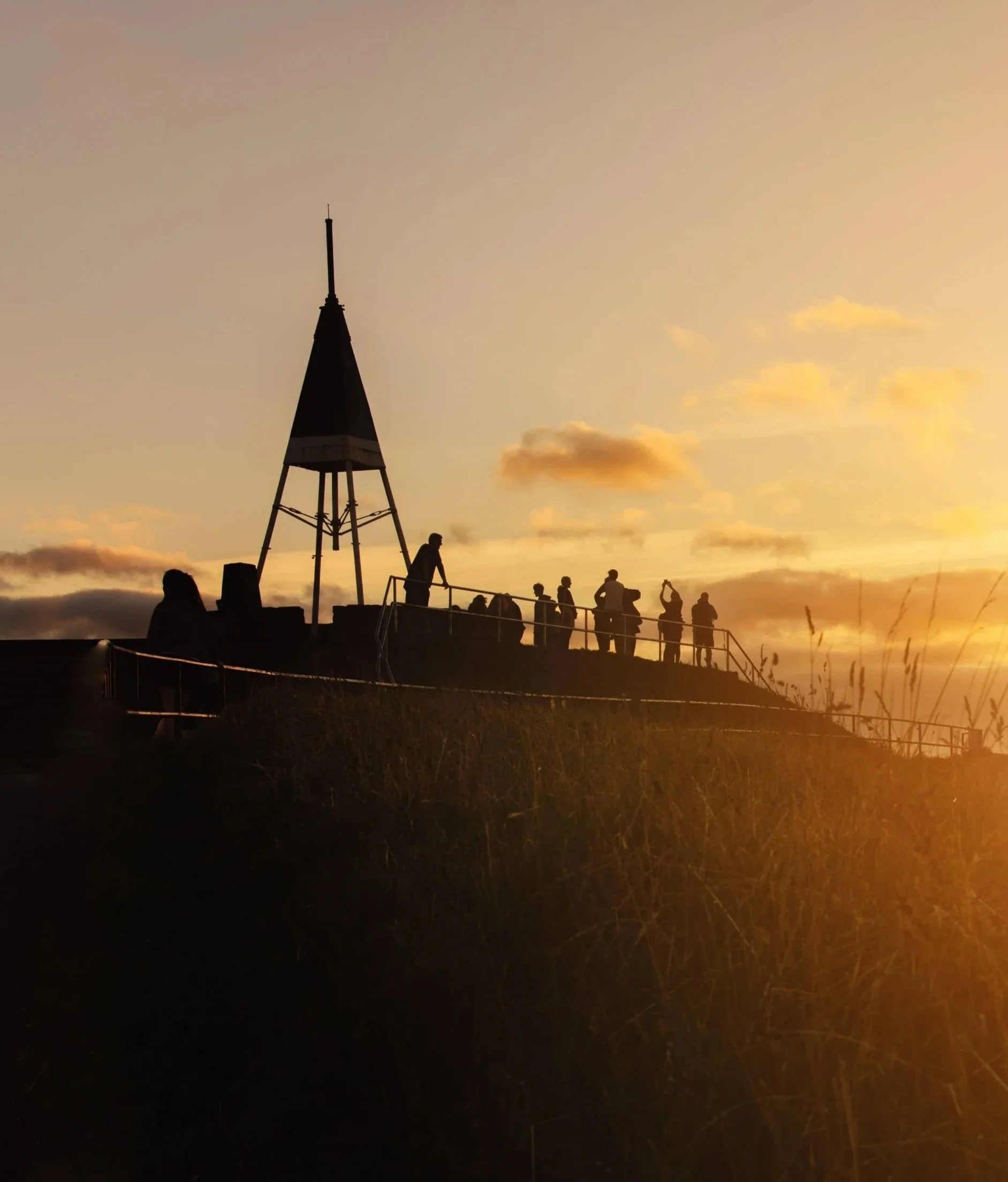 People standing on a hill at sunset with a wooden tower and clouds in the sky.