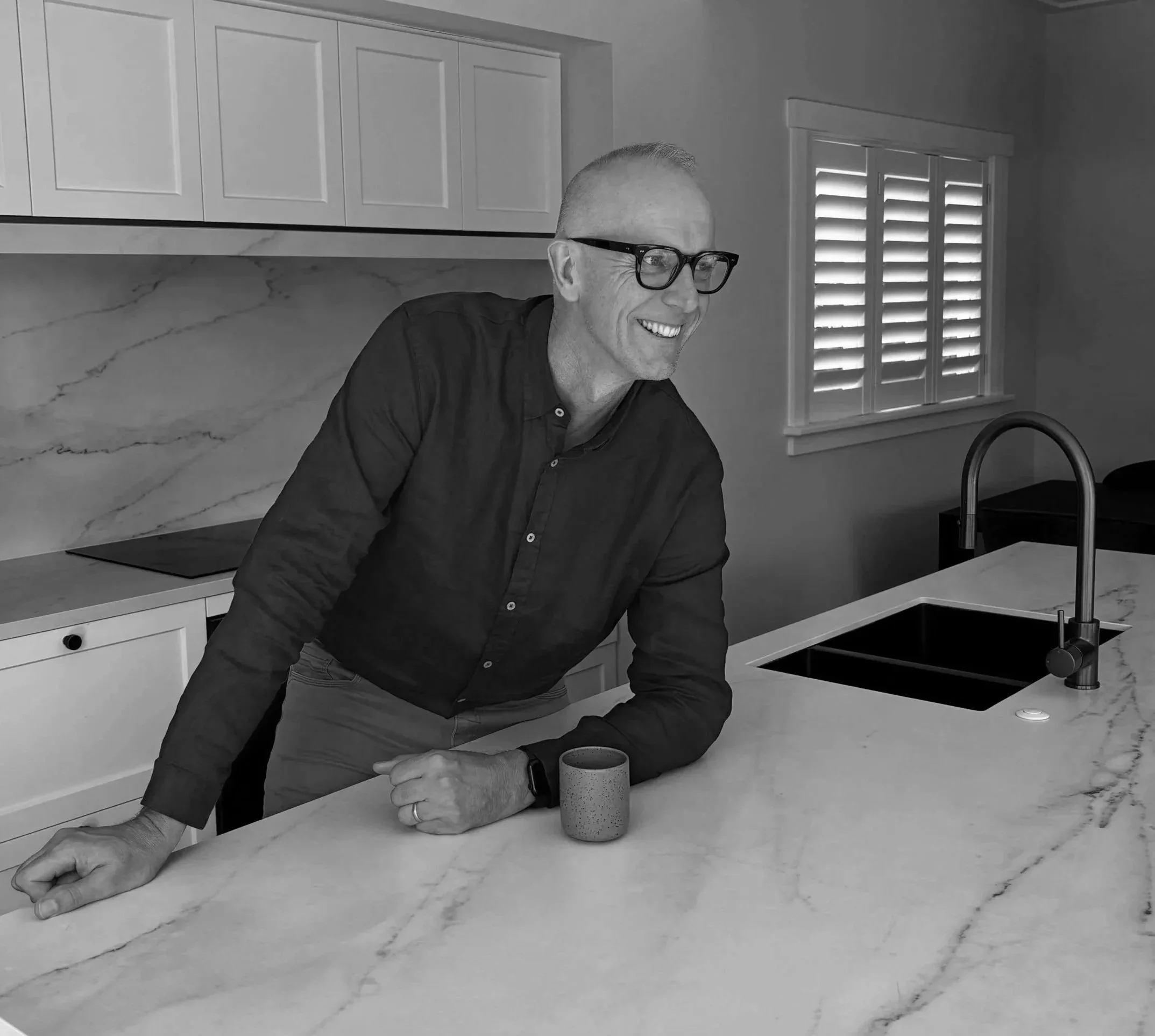 A man with glasses sitting at a kitchen counter, holding a cup with both hands, smiling, with kitchen cabinets and a window with shutters in the background.