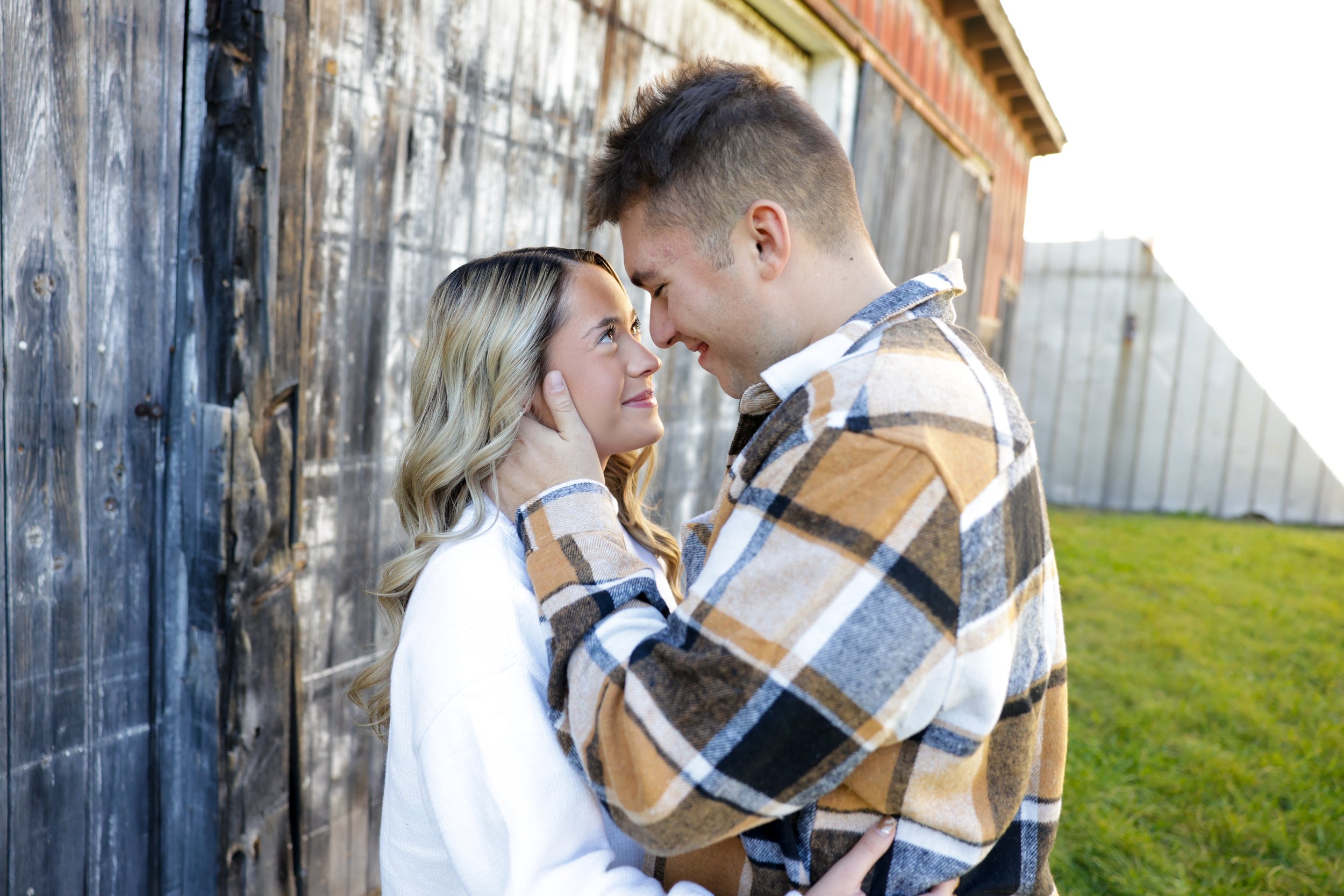 A Rochester couple standing close together outdoors, touching foreheads and smiling, with a wooden fence and a barn in the background in Pittsford, NY Canal
