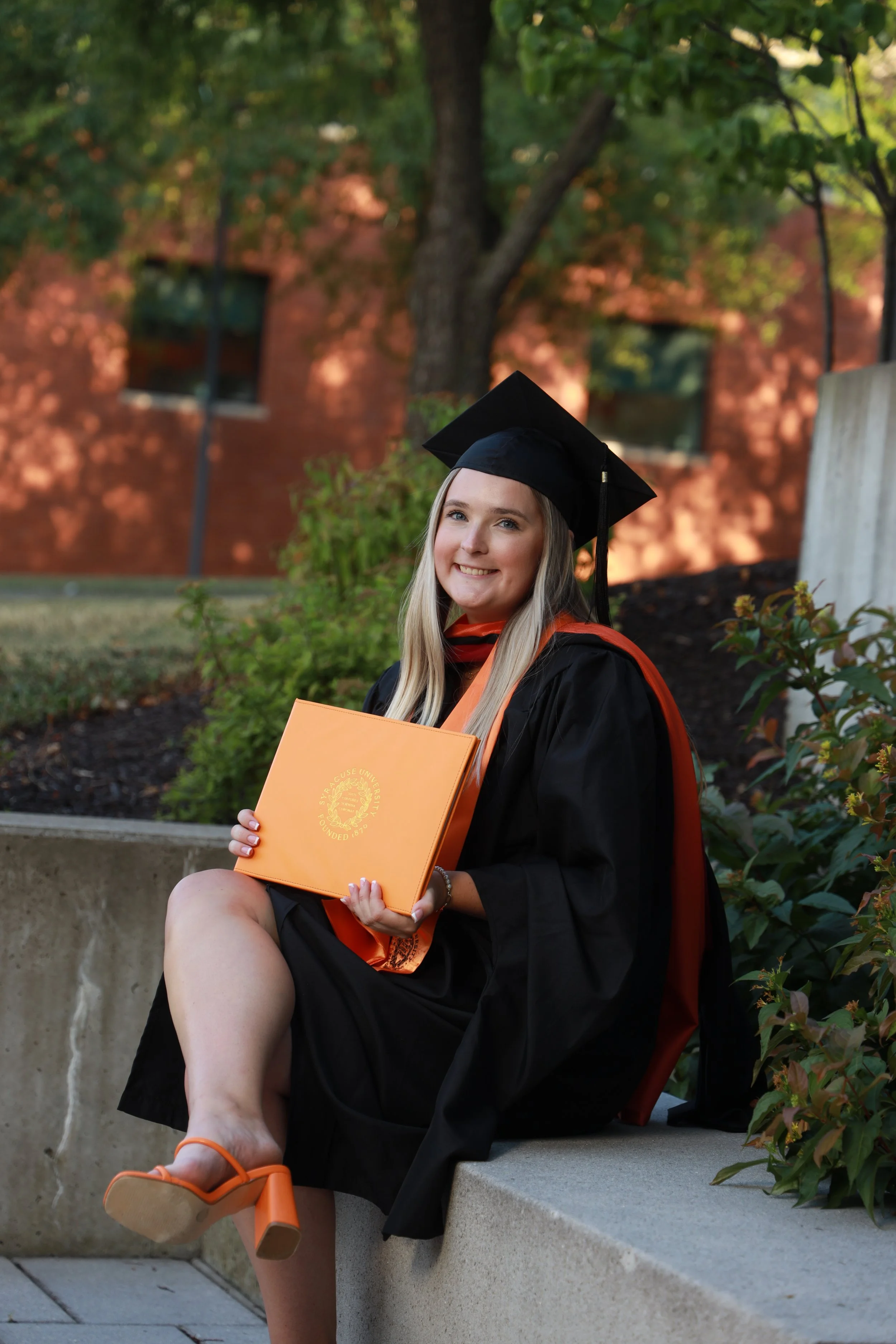 Young woman in graduation cap and gown sitting outdoors, holding a diploma, smiling.