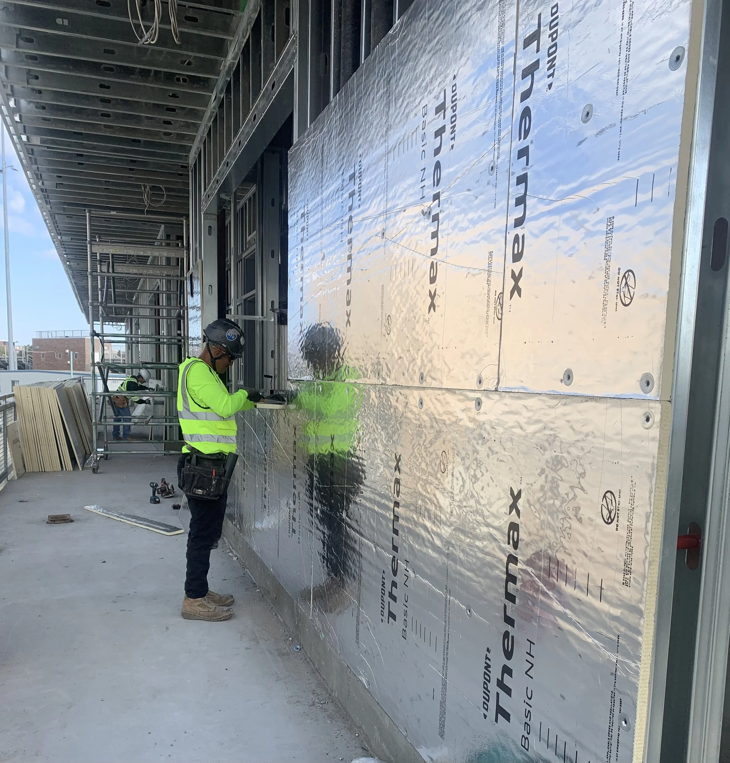 Construction worker in a yellow safety vest and helmet installing reflective insulation panels on a building exterior, with another worker in background.