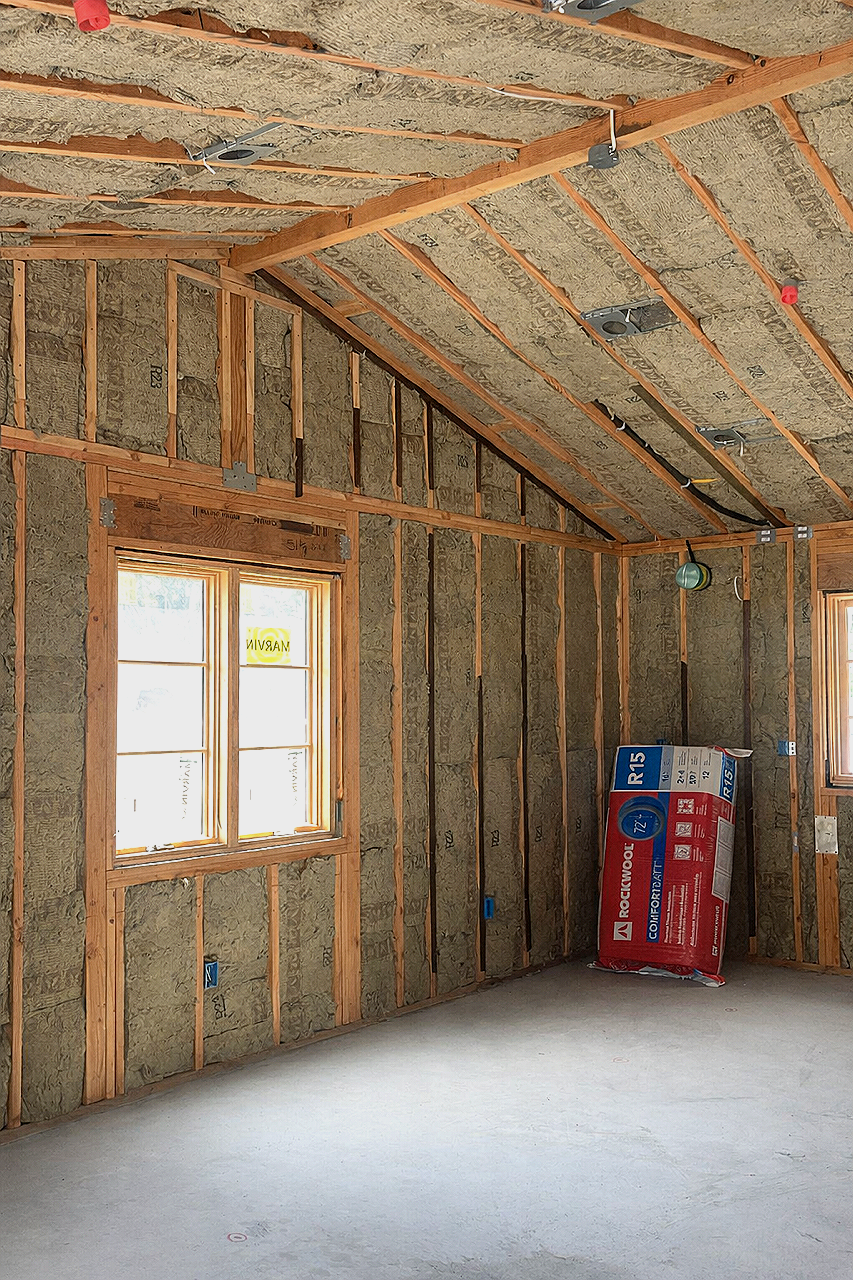 Shot of an unfinished room with insulation installed between the wooden wall studs and ceiling beams, two double-pane windows, and a bag of R-15 insulation in the corner.