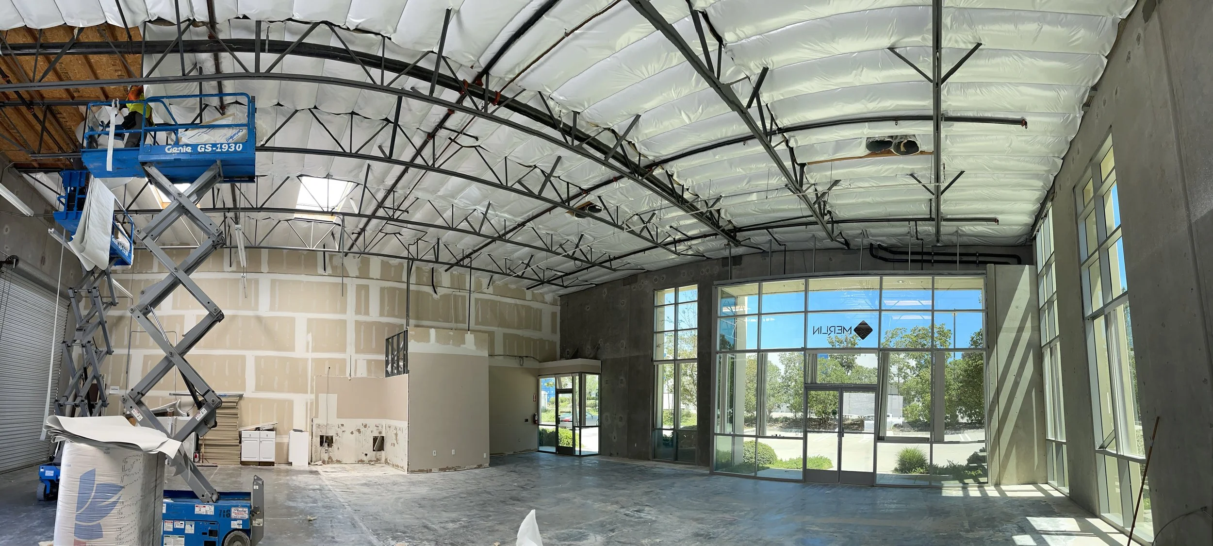 Interior of a commercial building under construction with large glass windows, a high ceiling with exposed insulation and structural elements, and a scissor lift on the left side.