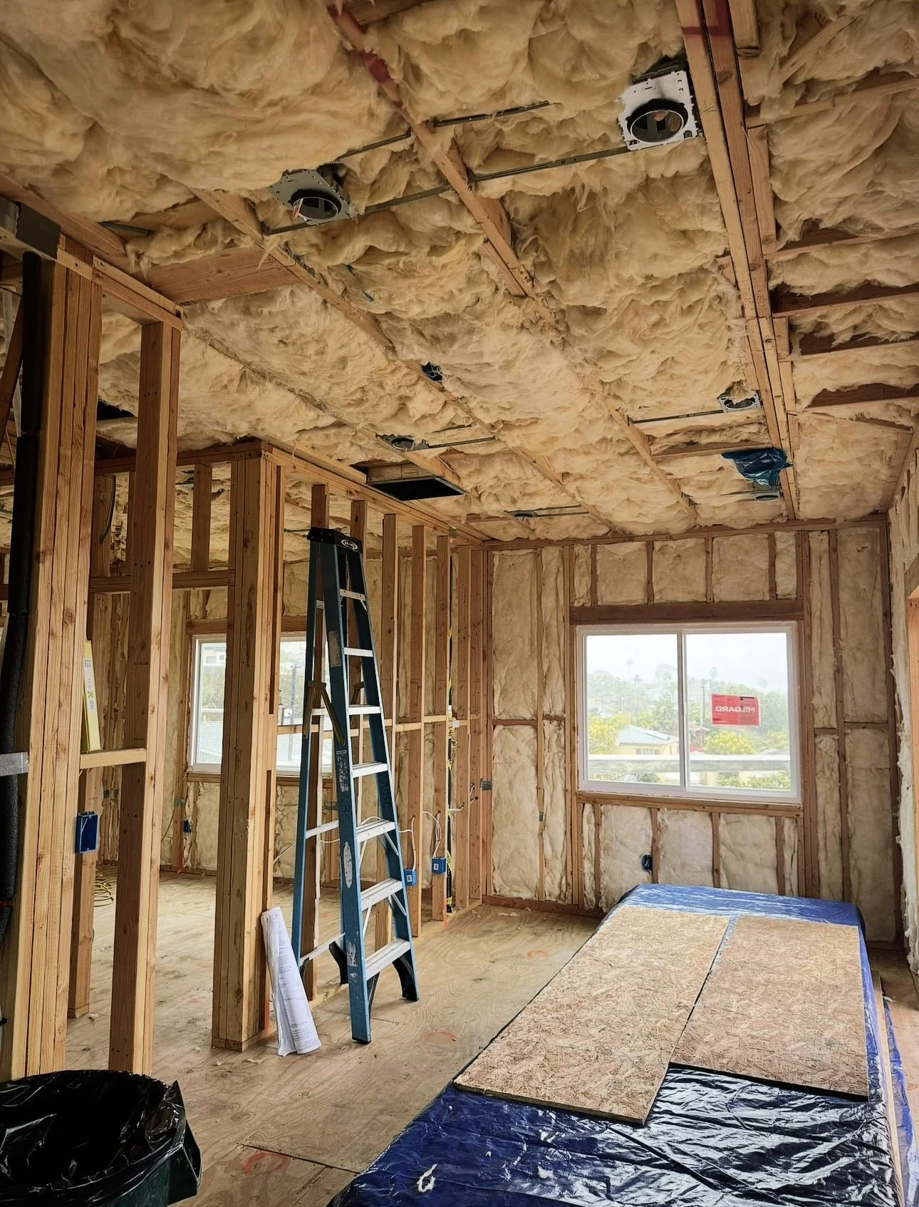 Interior of a room under construction with exposed wooden framing and insulation in the walls and ceiling. A blue ladder sits near the wall, and a window shows a view of trees and a cloudy sky outside. Construction materials and insulation covers are on the floor.