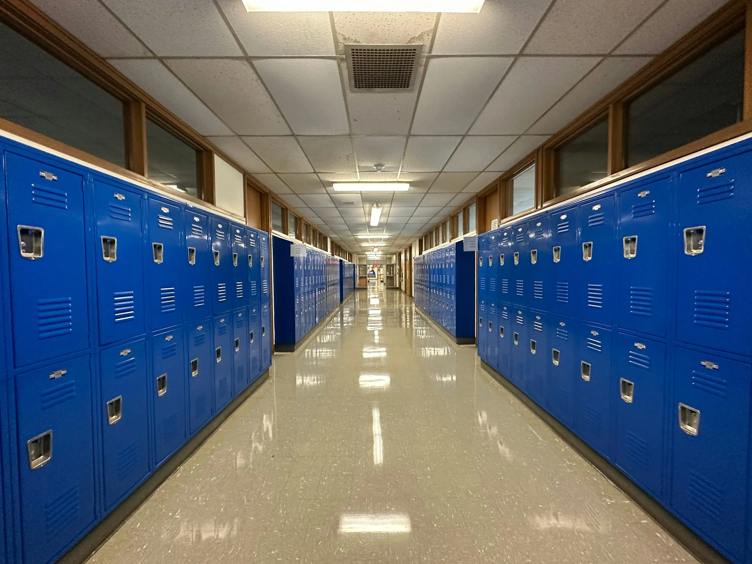 School hallway with blue lockers on both sides and windows above the lockers, leading to a distant exit door.