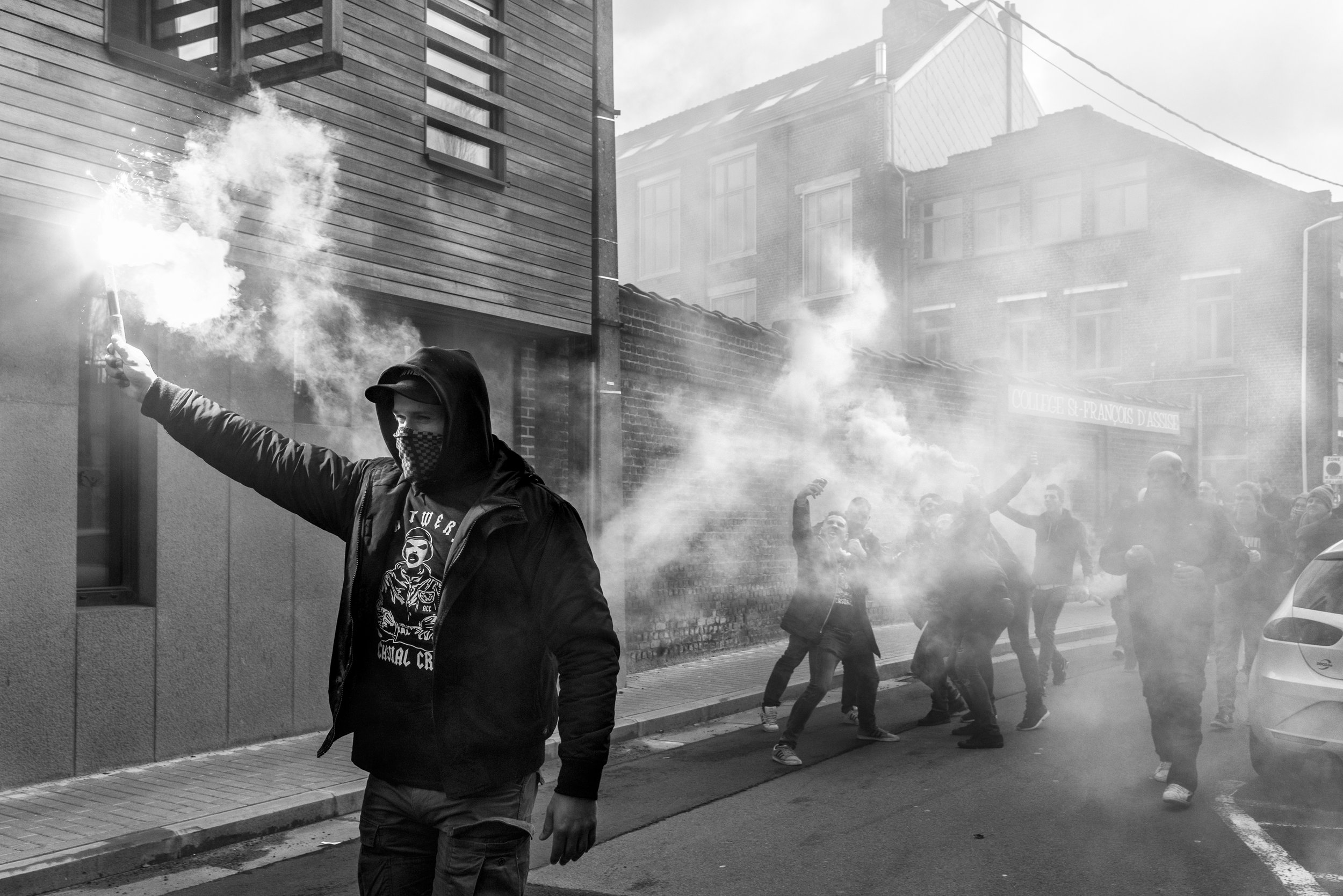 Een persoon met een capuchon en gezichtsmasker terwijl hij een rookbom vasthoudt in een straat, met een groep mensen op de achtergrond die alsof in protest of feestdrukte betrokken is, omgeven door rook.