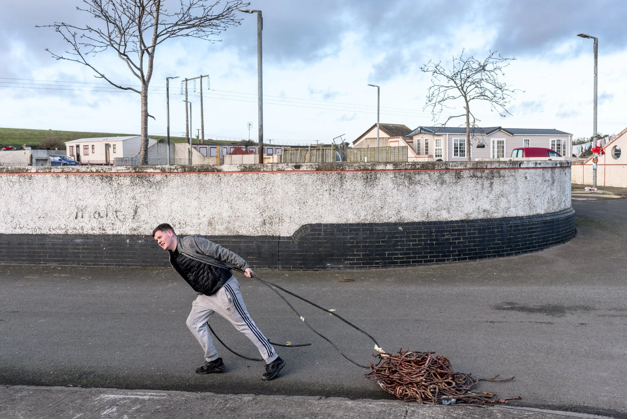 Een jongen die op de straat lijkt te lopen terwijl hij een bundel takken of vuilnis achter zich aantrekt, met een muur, bomen, en huizen op de achtergrond onder een bewolkte hemel.