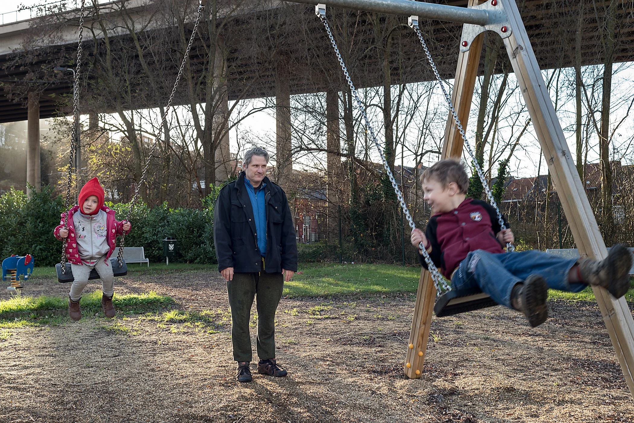 Twee kinderen op een speelplein, één op een schommel en de ander die in beweging is, terwijl een man ernaast staat en toekijkt.