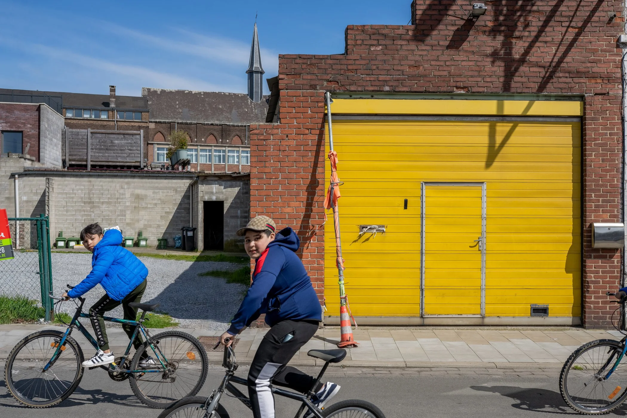 Twee jonge jongens fietsen langs een straat met een gele garage en een bakstenen gebouw op de achtergrond, onder een blauwe hemel.