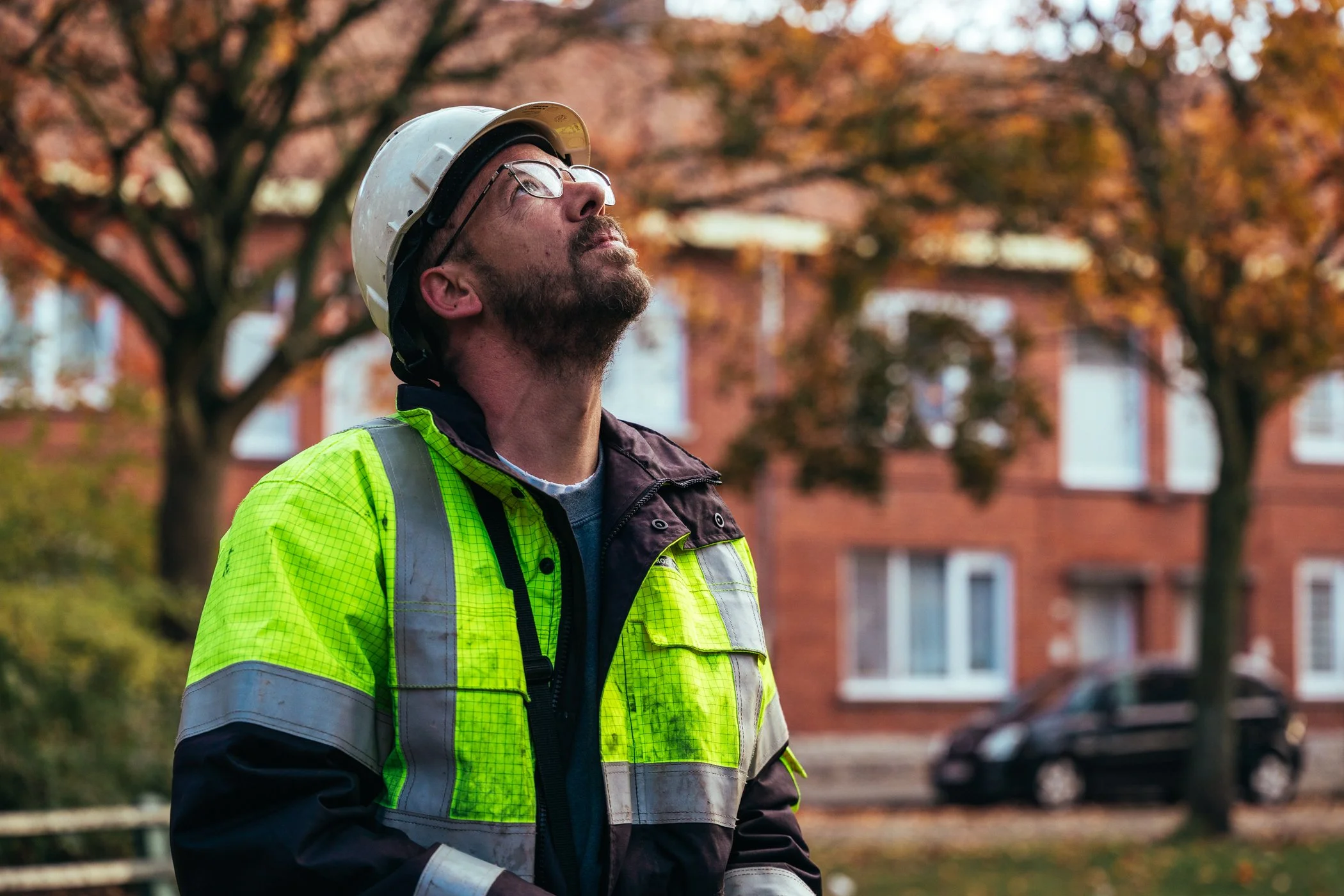 Man in arbeidskleding, een helm en veiligheidshesje, kijkt omhoog met een serieuze uitdrukking, terwijl hij buiten staat met bomen en een gebouw op achtergrond.