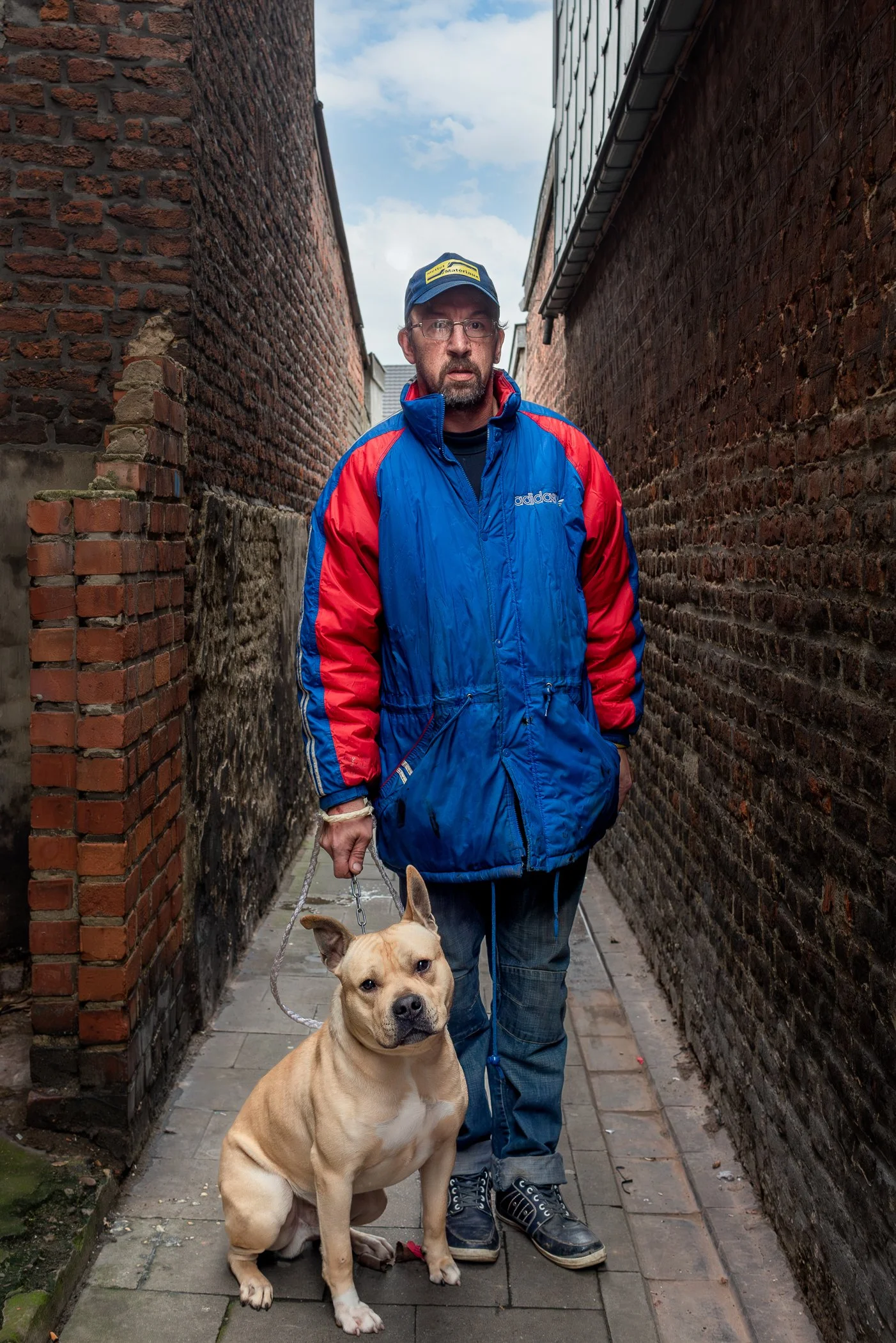 Man in een blauwe en rode jas met een hond in een smalle steeg tussen bakstenen muren, bewolkte hemel zichtbaar.