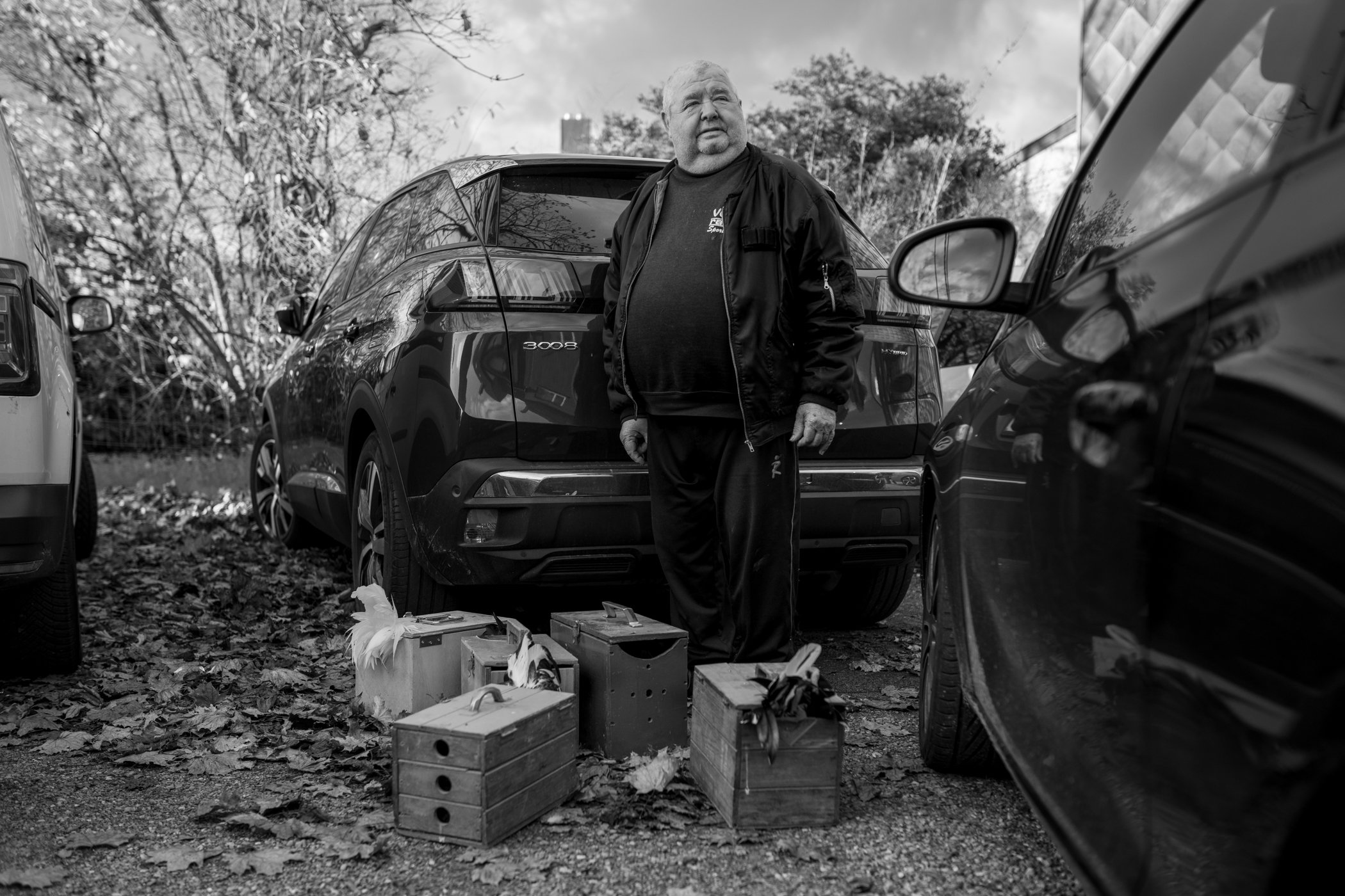 Een man in een zwarte jack staat tussen geparkeerde auto's, met meerdere houten dozen op de grond, onder een gedeeltelijk bewolkte hemel.