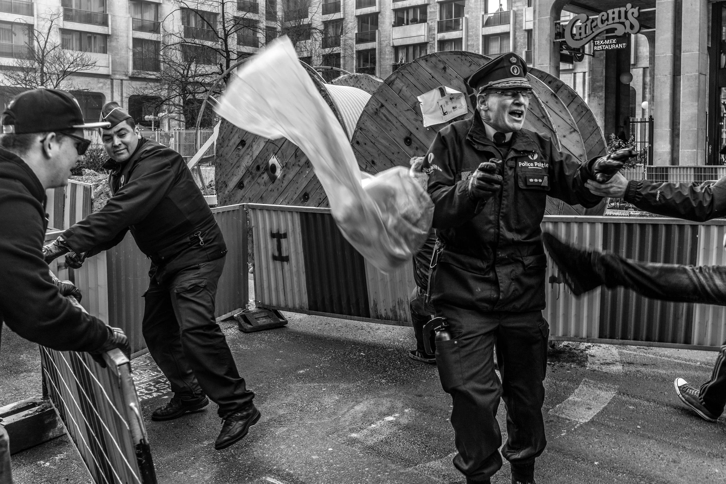 Gezicht van een politieagent die zich verzet tijdens een protest of demonstratie, omringd door barricades en andere personen in een stedelijke omgeving.