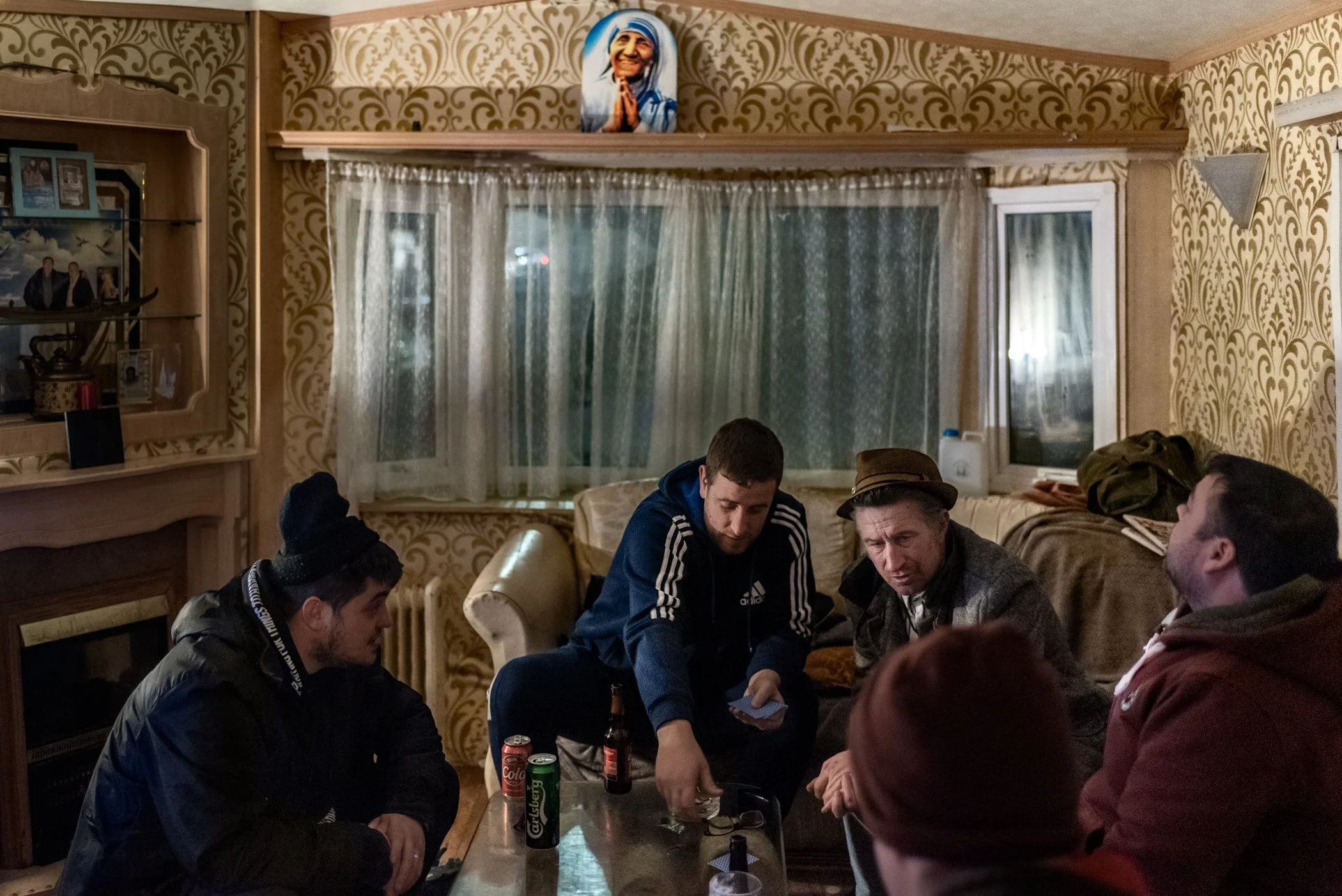 Vijf mannen zitten binnen in een woonkamer rond een tafel, die speelt met kaarten. Op de tafel staan verschillende drankjes, waaronder cola en bier. Achter hen is een raam met witte transparante gordijnen, en aan de muur hangt een afbeelding van een 