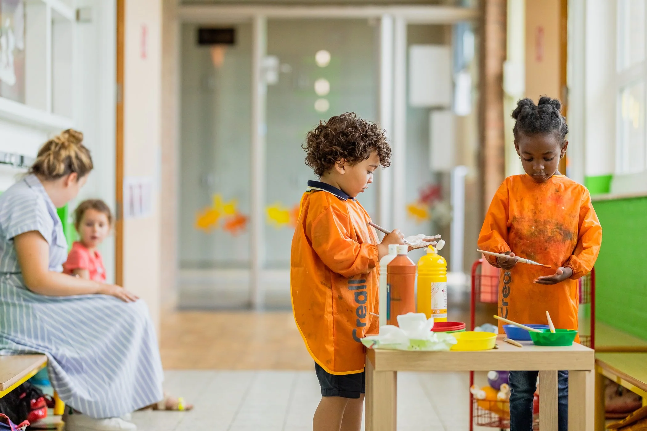 Twee jonge kinderen, een jongen en een meisje, zitten aan een kleine tafel en lijken te schilderen met verf. Ze dragen oranje schildersmantels. In de achtergrond zitten twee andere kinderen op een bank, een volwassene met een jurk en een baby.