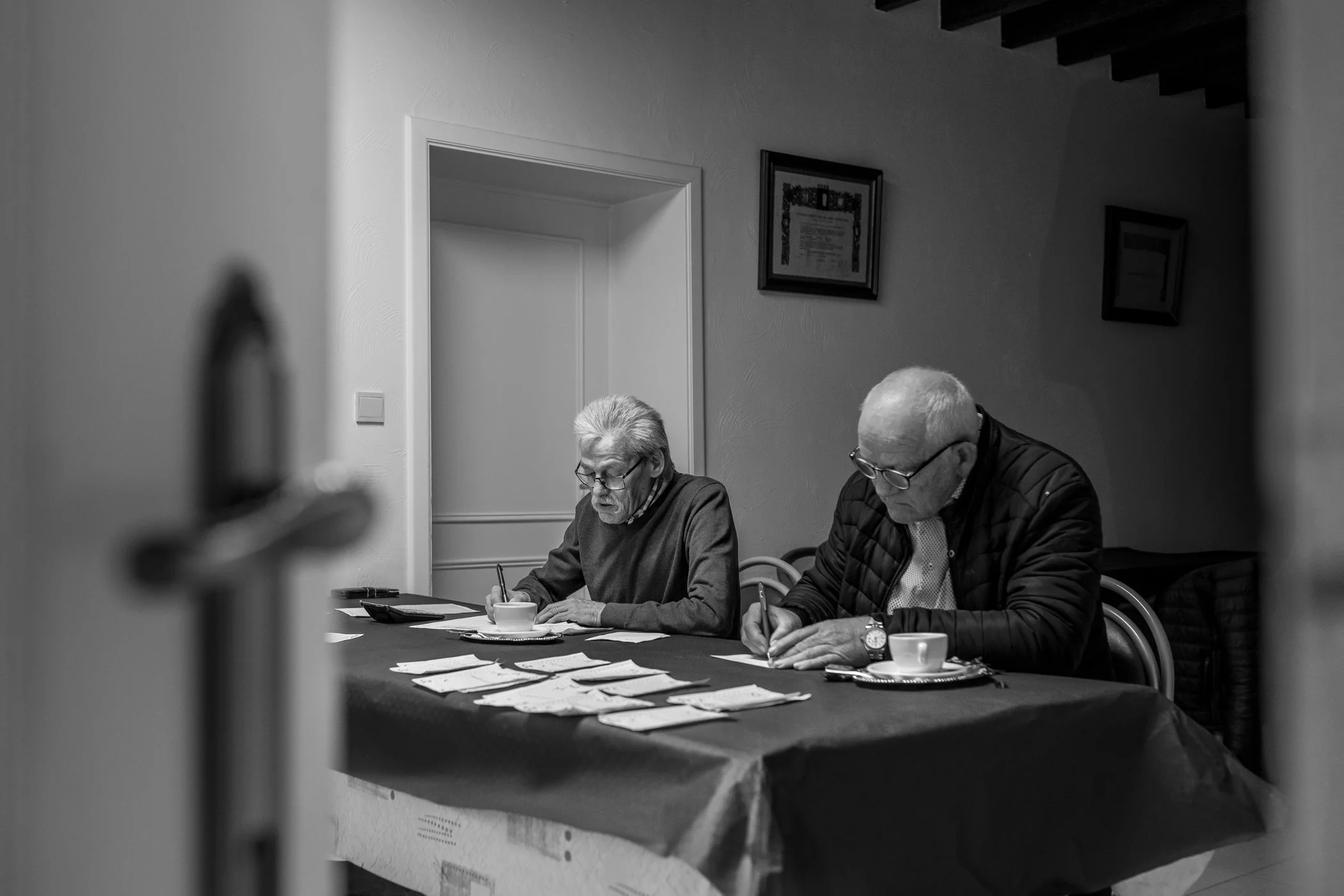 Twee oudere mensen zitten aan tafel, een man en een vrouw, met koffie en papieren, ze schrijven en kijken geconcentreerd.