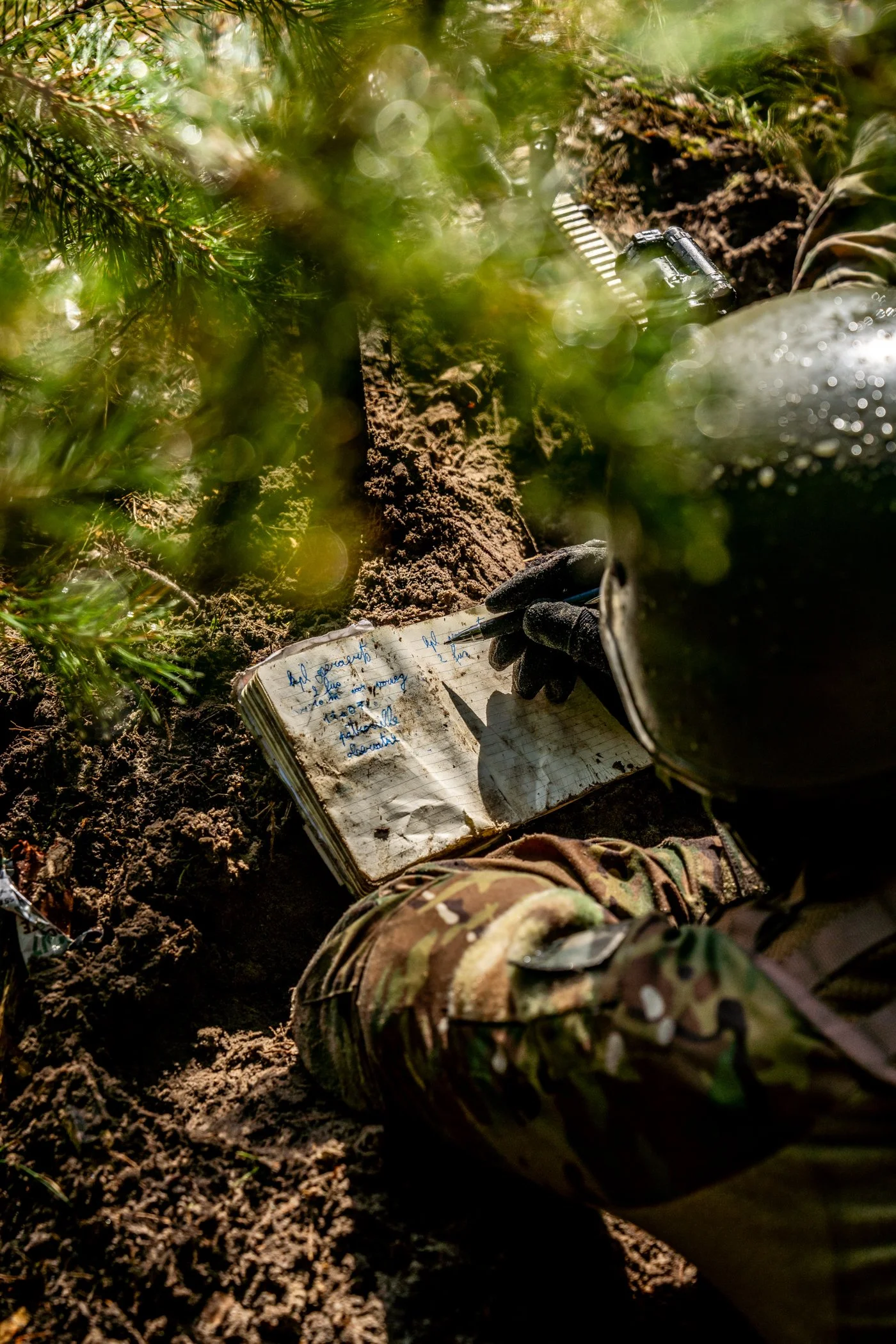 Een militair die in de modder op de grond zit, een notitieboekje vasthoudt en aantekeningen schrijft. De militair draagt camouflage-uitrusting en een helm, met omringende groene struiken en aarde.