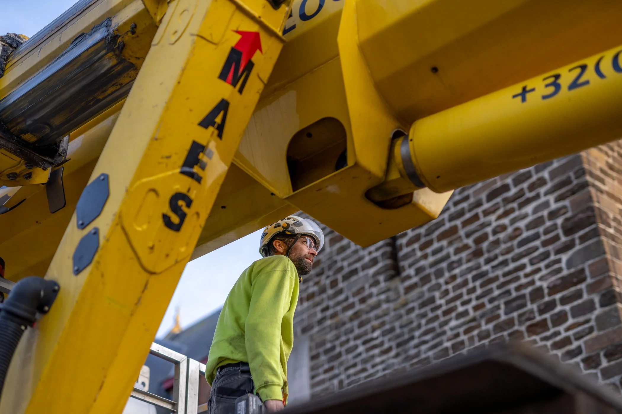 Een bouwvakker met helm en veiligheidsbril staat onder een grote gele ladder of kraan, voor een bakstenen muur.