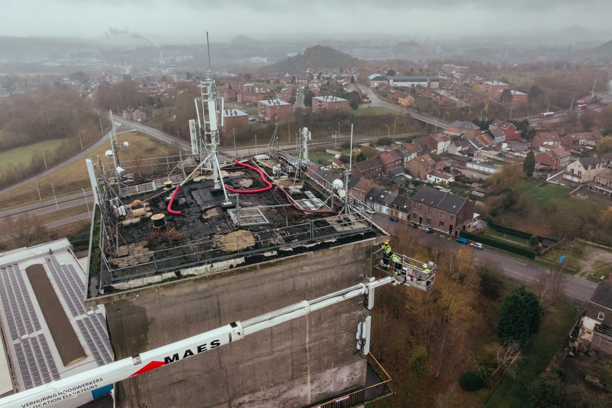 Uitzicht vanaf hoogte van een hoog gebouw met brandplekken op het dak, brandweerlieden op een héli-platform, terrassendaken, huiswijken en wegen onder een bewolkte hemel.