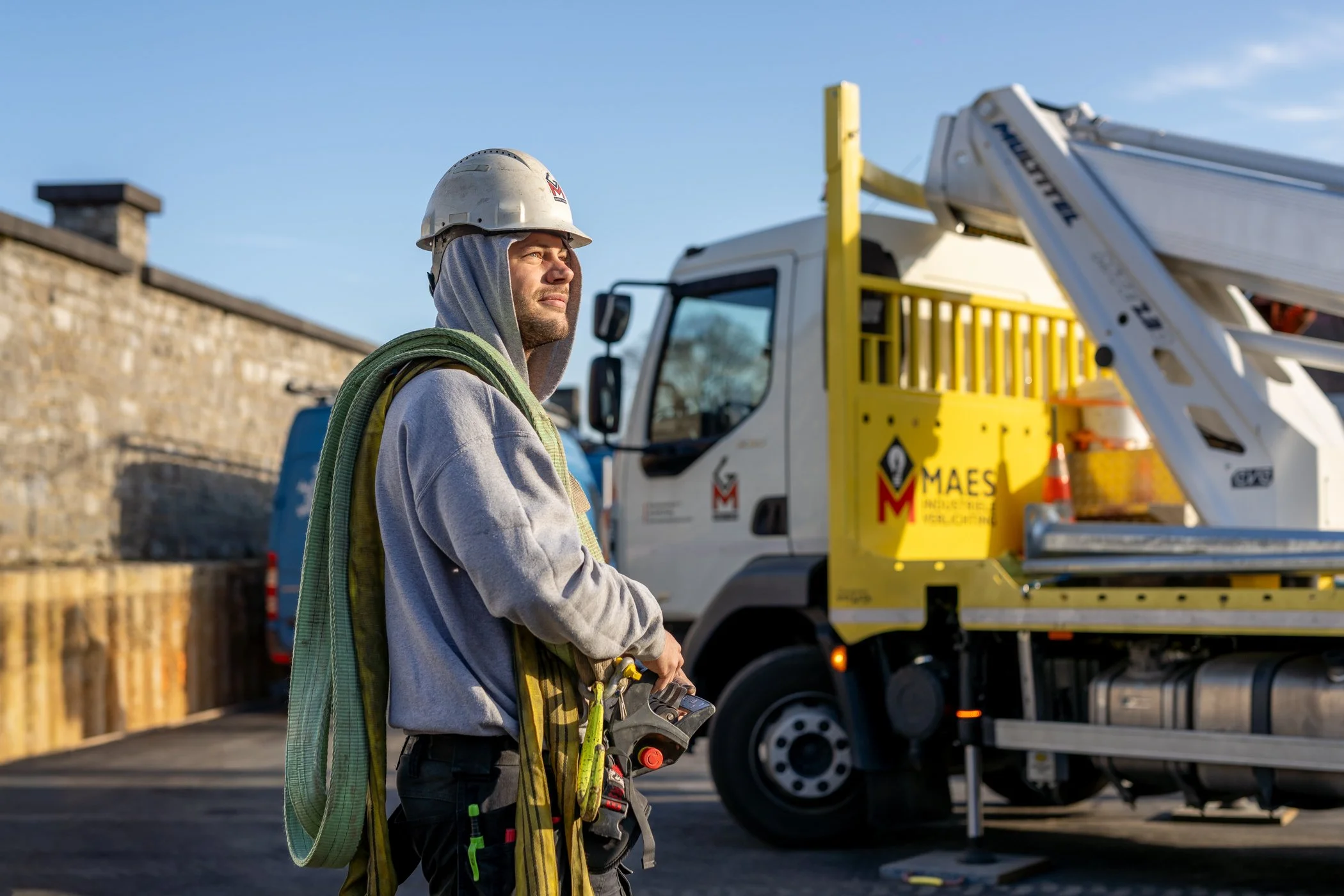 Een man in bouwhelmp en werkoutfit staat naast een werkwagen op een bouwplaats, met een neerslag van de zon op zijn gezicht.