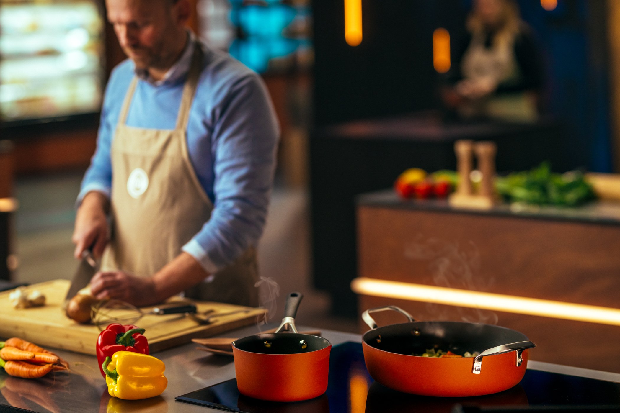 Een man kookt in een keuken met kappen met groenten en piment in de voorgrond.
