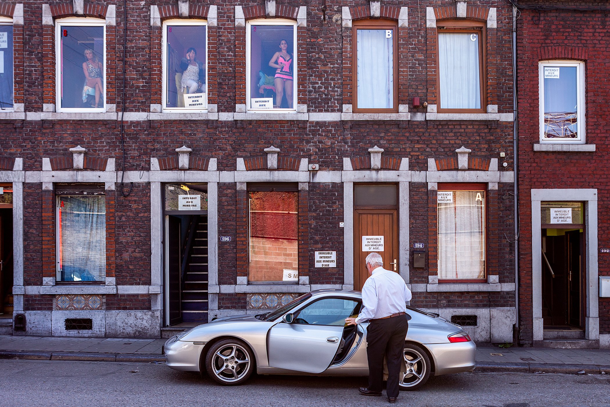 Een man met witte haar en witte overhemd staat naast een zilverkleurige auto op straat, voor een gebouw met ramen en ramen met mannequins draperieën, en verschillende waarschuwings- en verbodenborden.