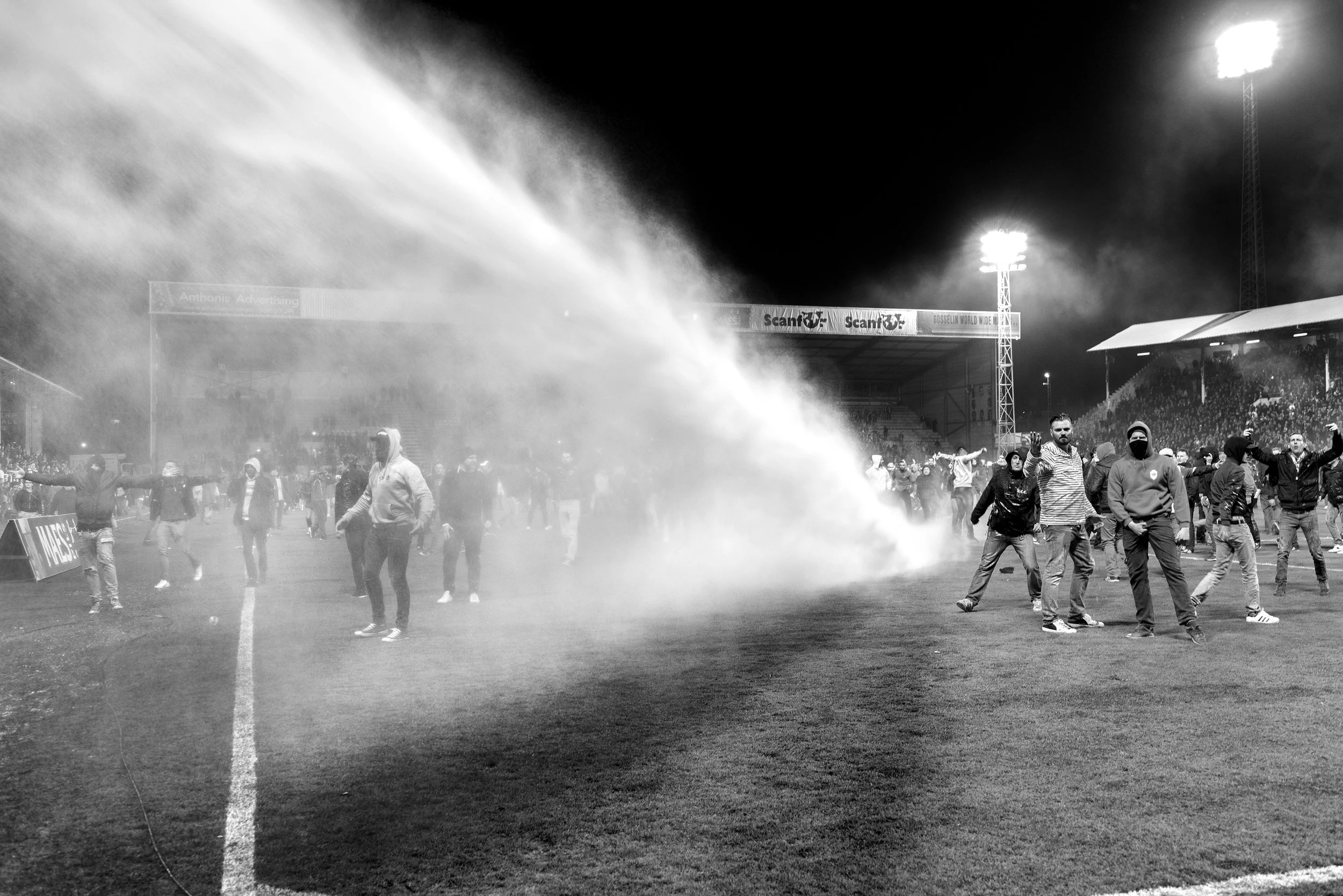 Mensen op een voetbalstadion veld tijdens een protest of rellen, met rook en tegenspelers op de achtergrond, in zwart-wit.