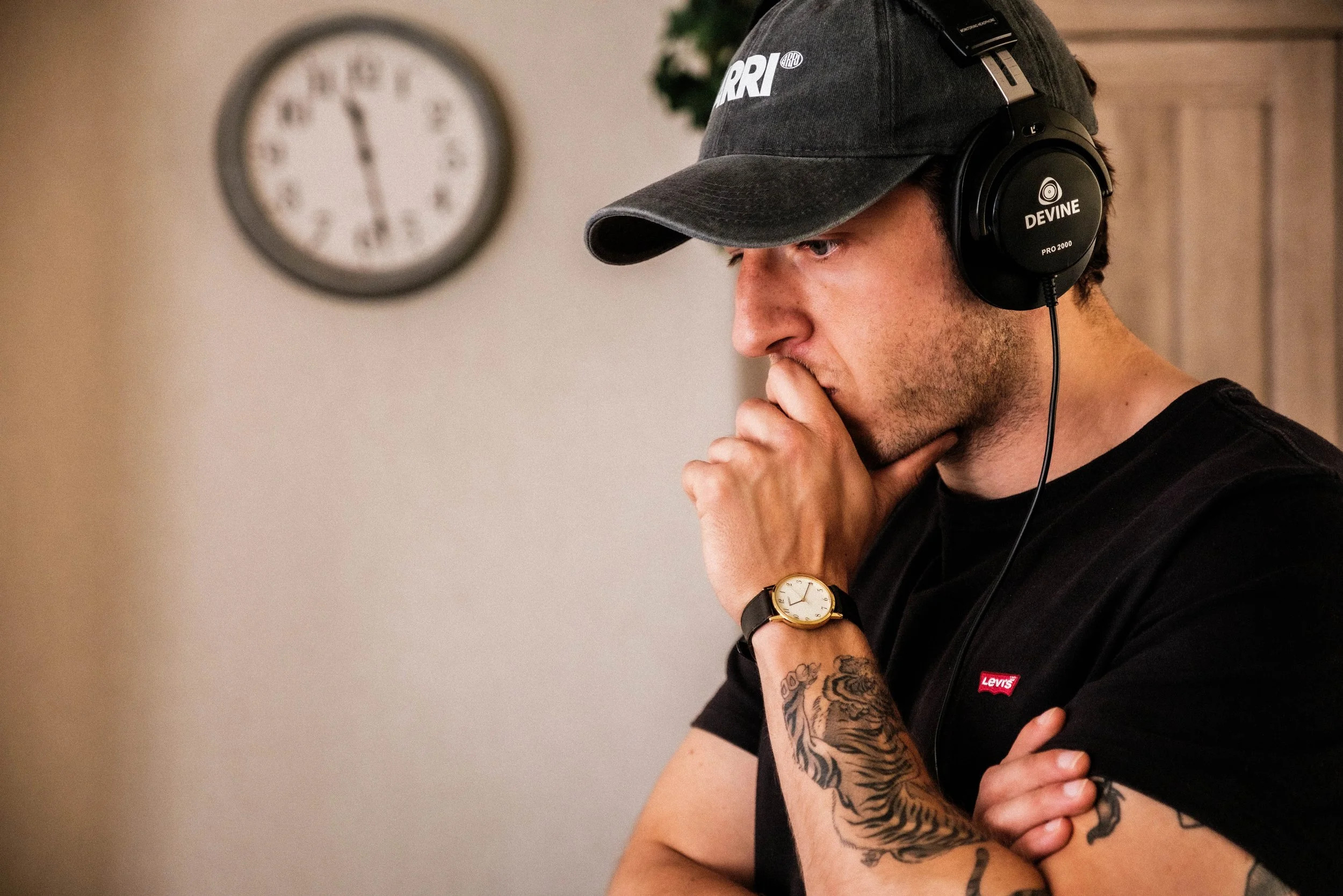 Man met een zwarte T-shirt en sporthorloge, draagt een zwarte cap en koptelefoon, denkt met hand op kin, in een kamer met een klok op de muur.