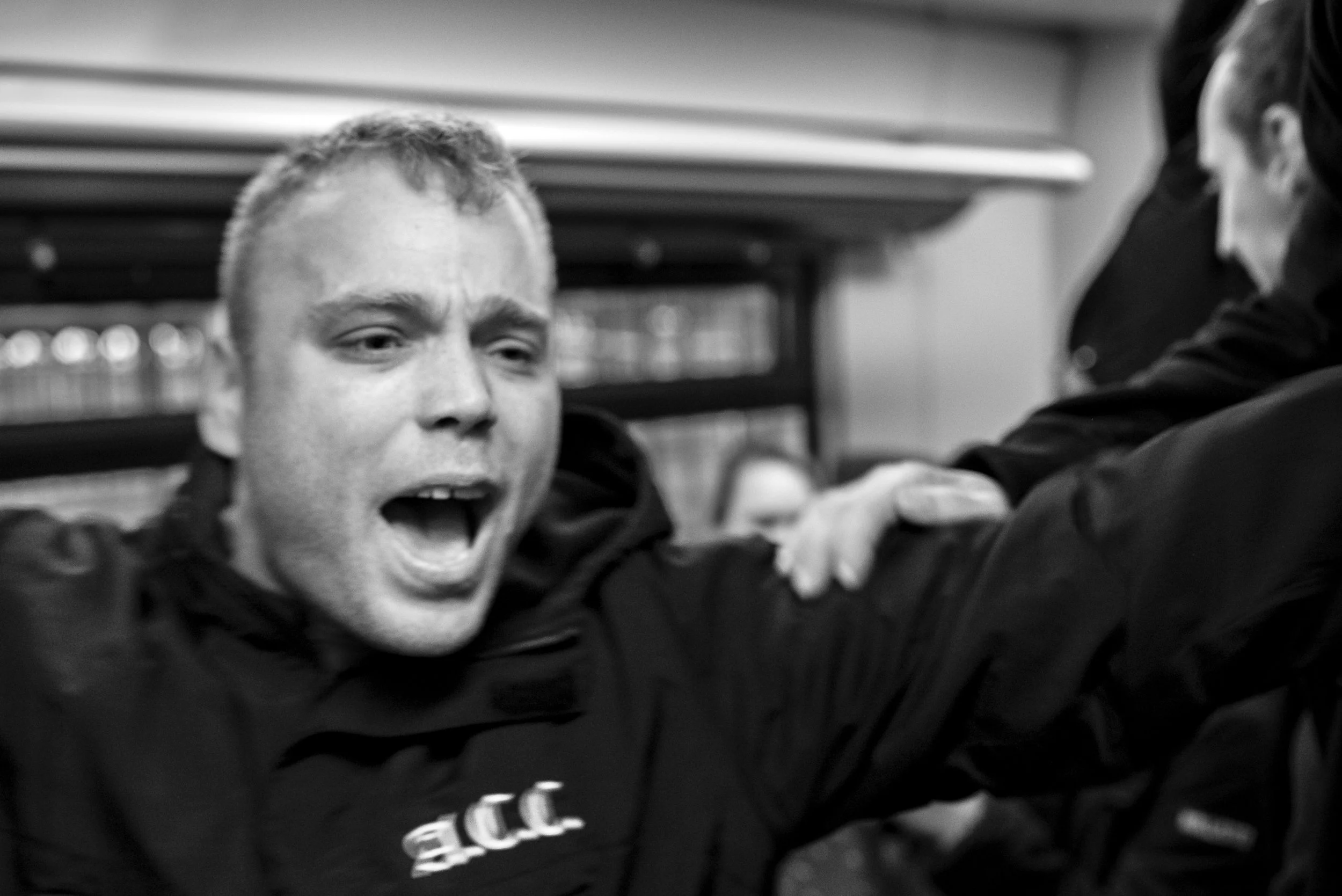 Een man met een korte baard en kort haar schreeuwt terwijl hij omringd is door anderen, op een sportschool of trainingslocatie.