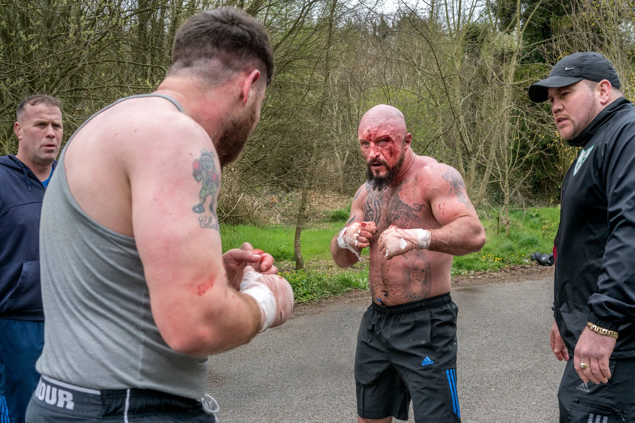 Twee getatoeëerde mannen en een man in een sportoutfit praten buiten in de natuur, met een andere man die zich voorbereidt op een wedstrijd.