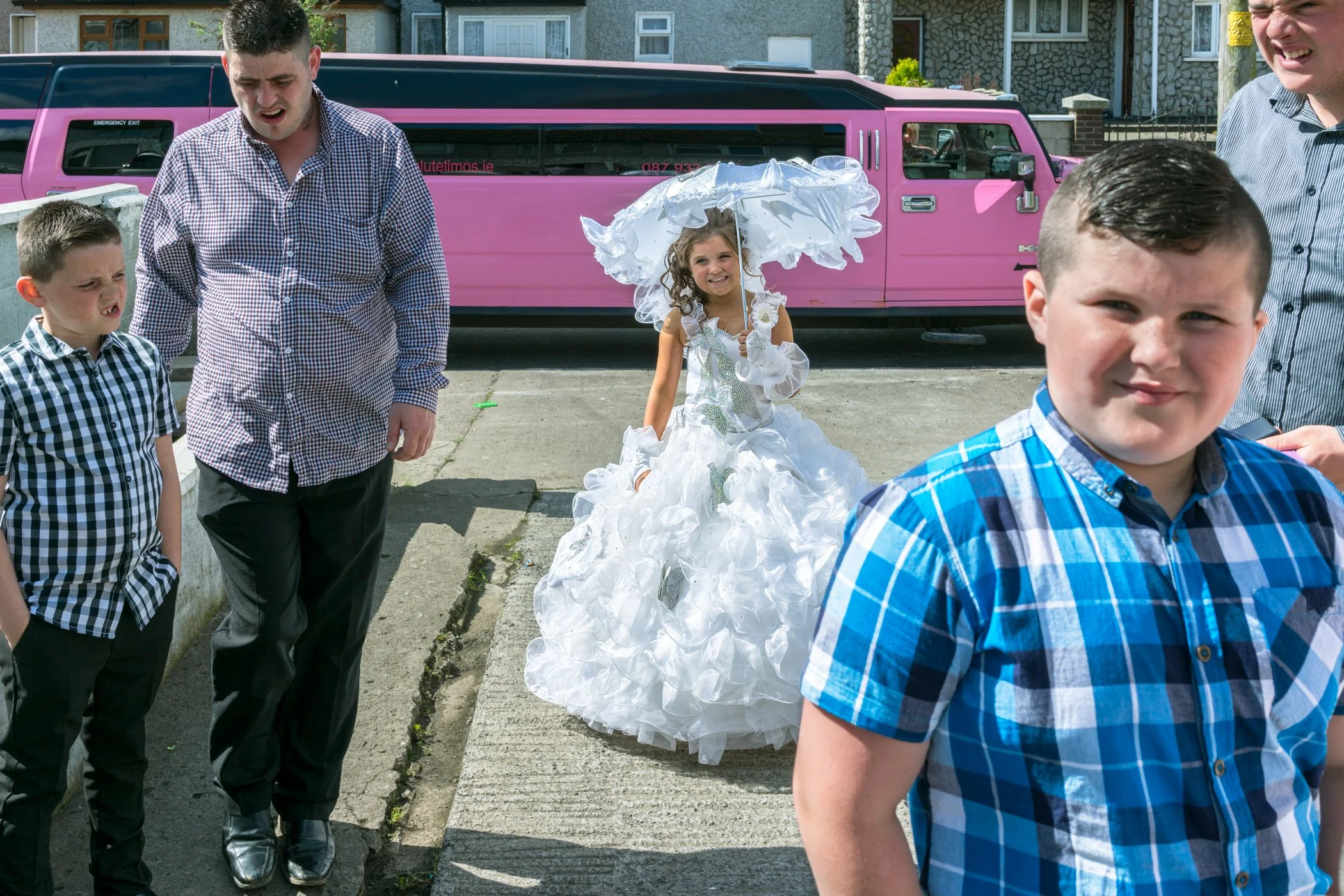 Een jonge meisje in een witte, bloemrijke jurk met een parasol staat op een trottoir, omringd door enkele jongens en mannen, buiten op straat met een roze limousine op de achtergrond.