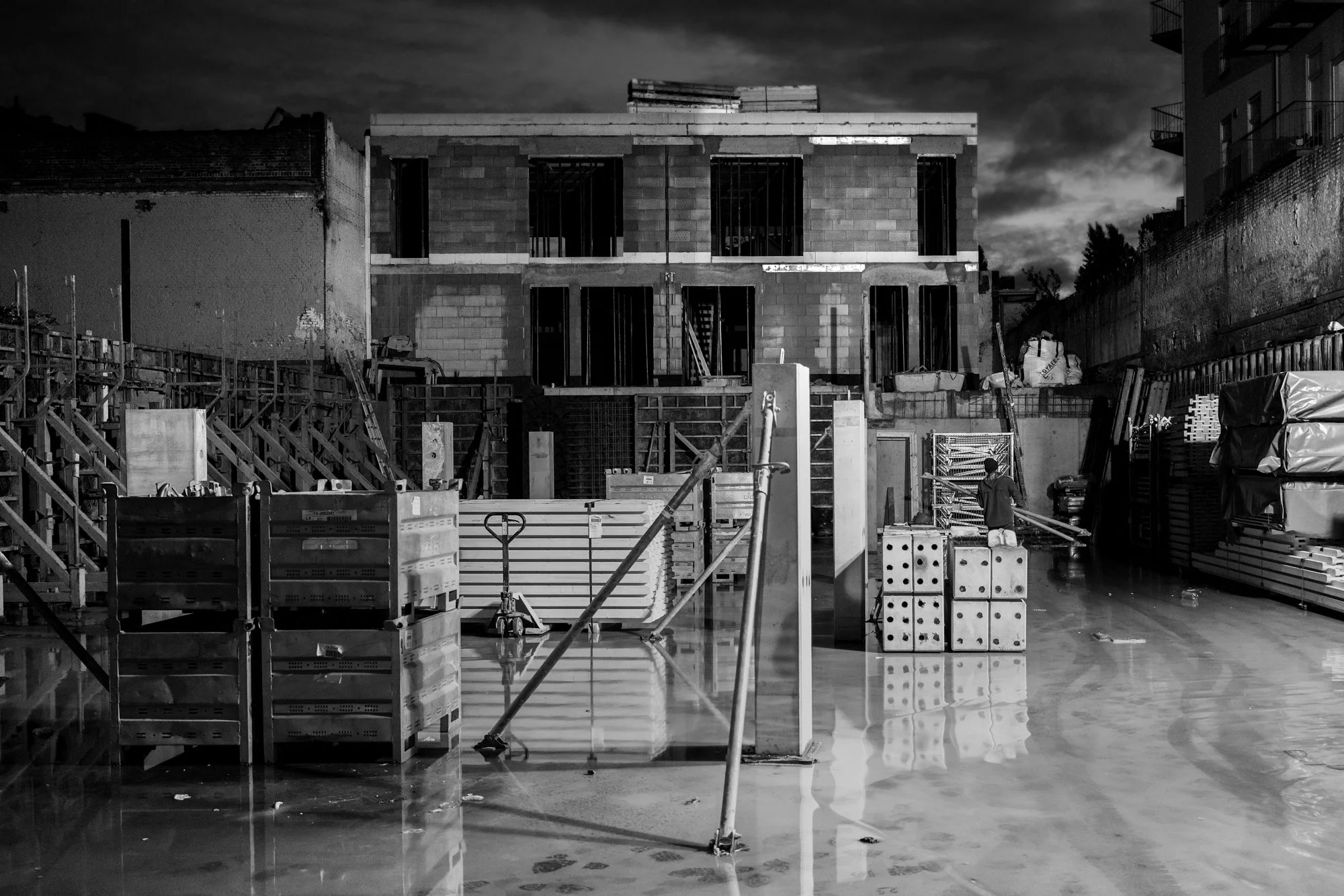 Een bouwplaats met een half afgewerkte structuur, bouwmaterialen en een man die lijkt te werken, onder een donker bewolkte hemel.