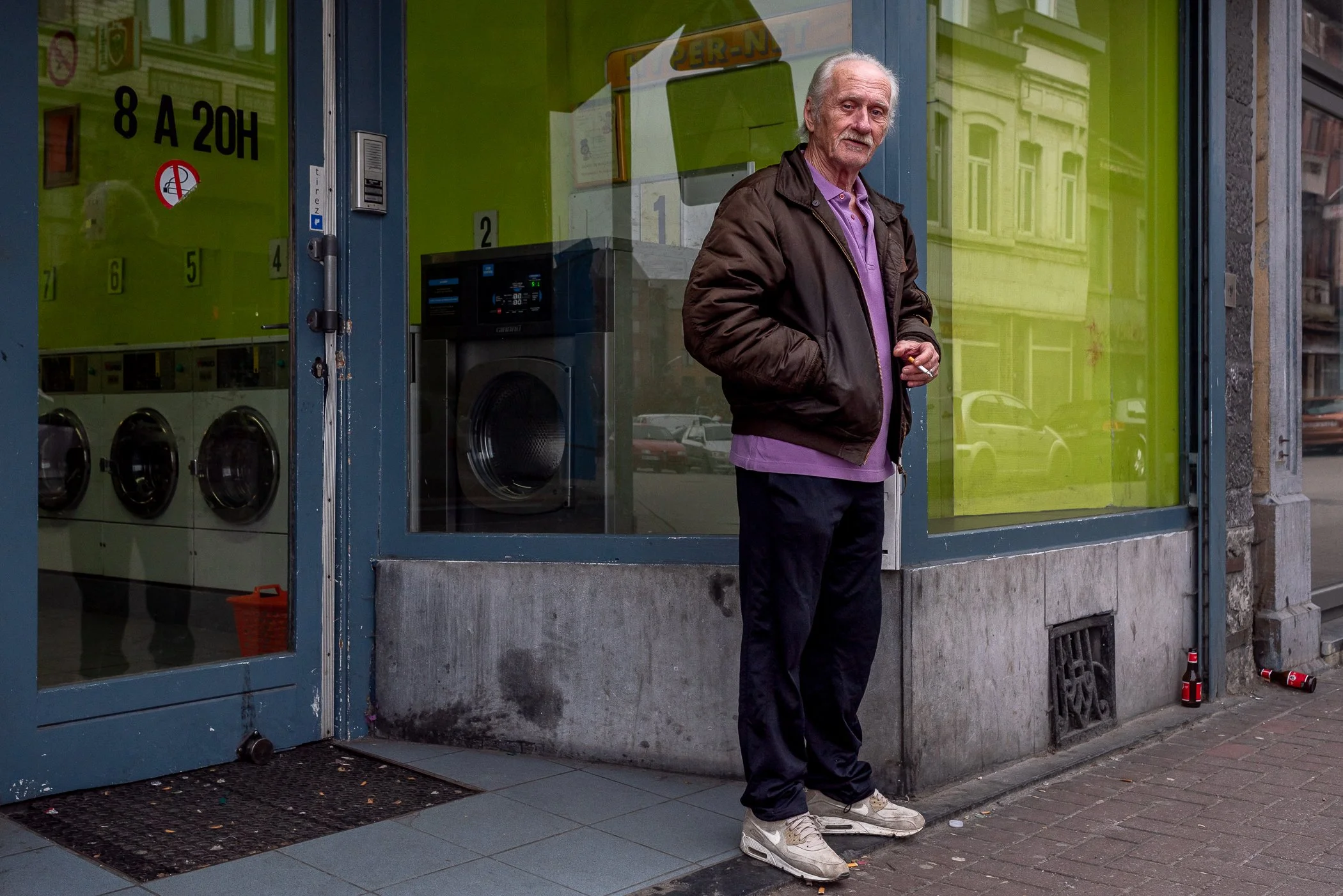 Een oudere man staat buiten een wasserette met een paar lege flesjes op de stoep. Hij draagt een donkerbruin jack, een paar witte sneakers en heeft grijs haar en een baard.