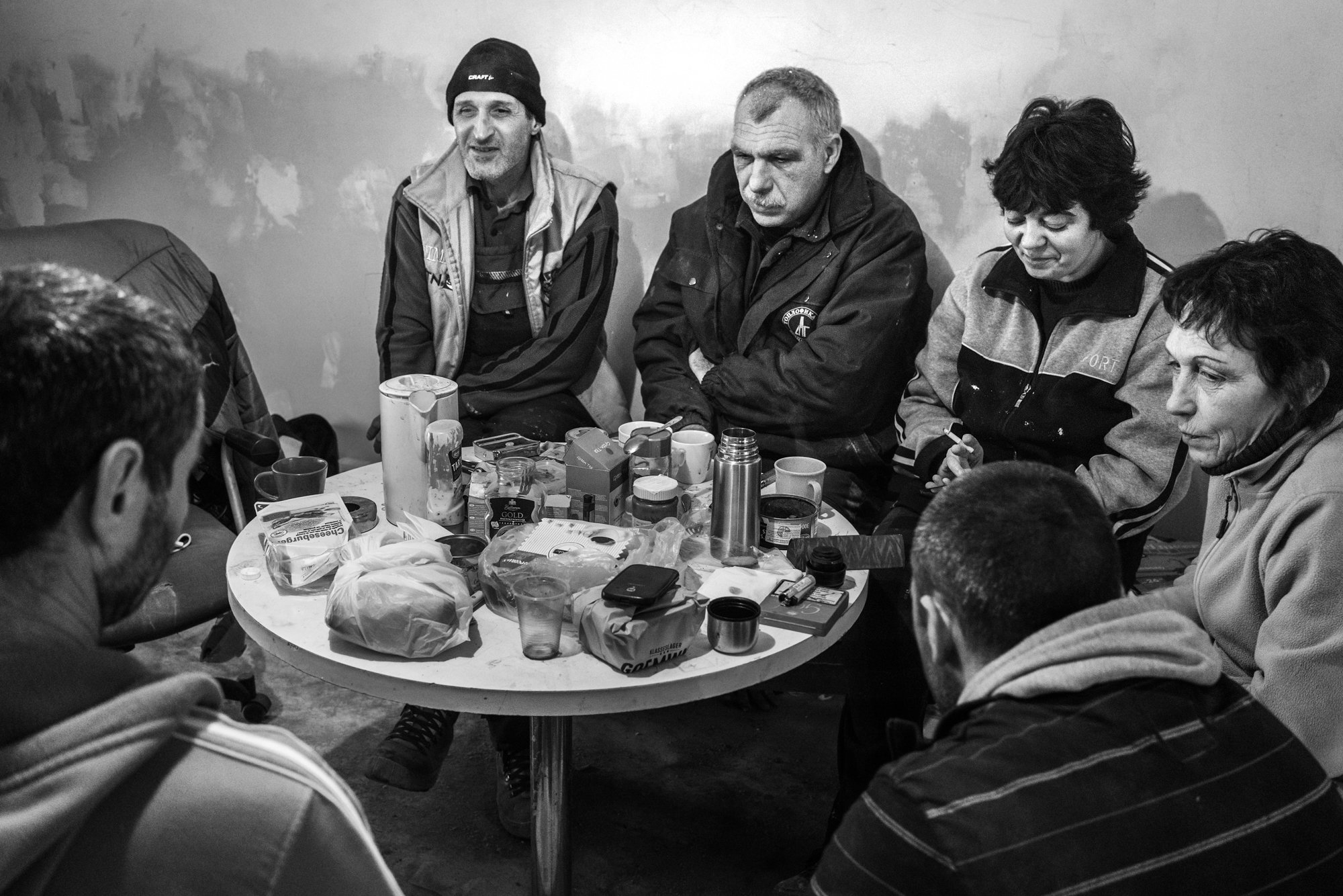 Zes mensen zitten rondom een tafel met voedsel en drinken, binnen een kamer met een onafgewerkte muur in zwart-wit.