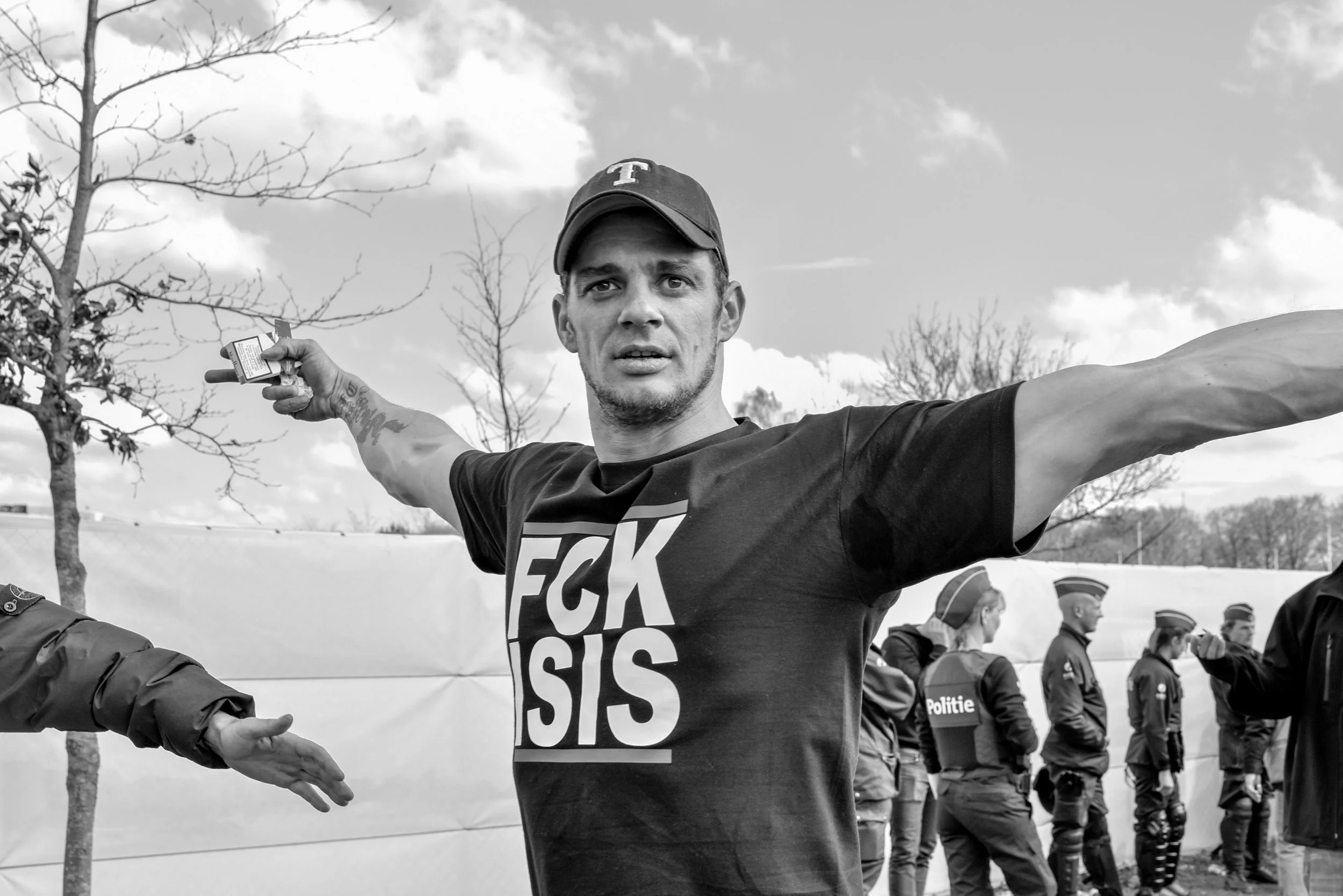 Een man met uitgestrekte armen, draag een zwart t-shirt met de woorden 'FCK ISIS' en een baseballcap, omringd door politieagenten tijdens een protest of demonstratie.