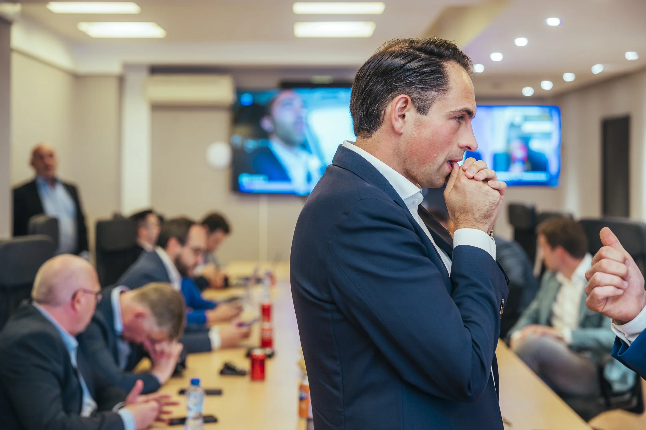 Man in een donkerblauw pak die nadenkt tijdens een vergaderingsvergadering met andere mensen aan een lange tafel.