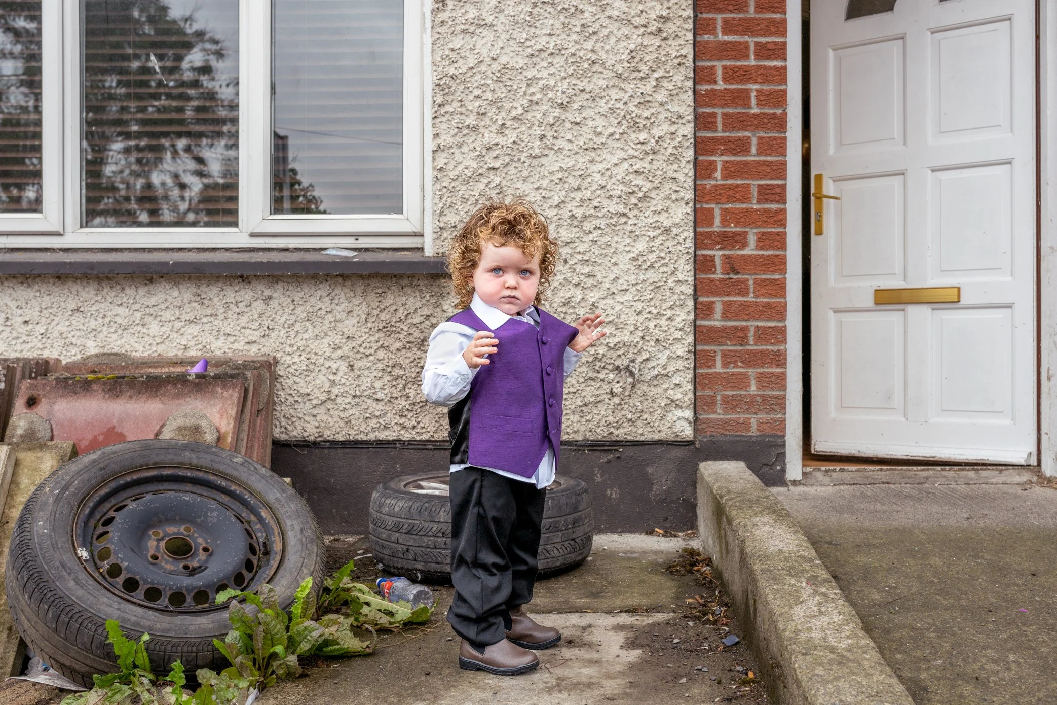 Jongen met krullend haar, gekleed in een wit overhemd en een paarse vest, staat buiten voor een huis met een deur en een raam, omringd door auto-velgen, afval en planten.